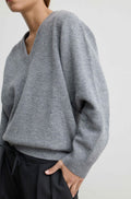 A close-up of a person wearing an oversized heather gray V-neck knit sweater with ribbed trim, paired with dark pants against a plain light background.
