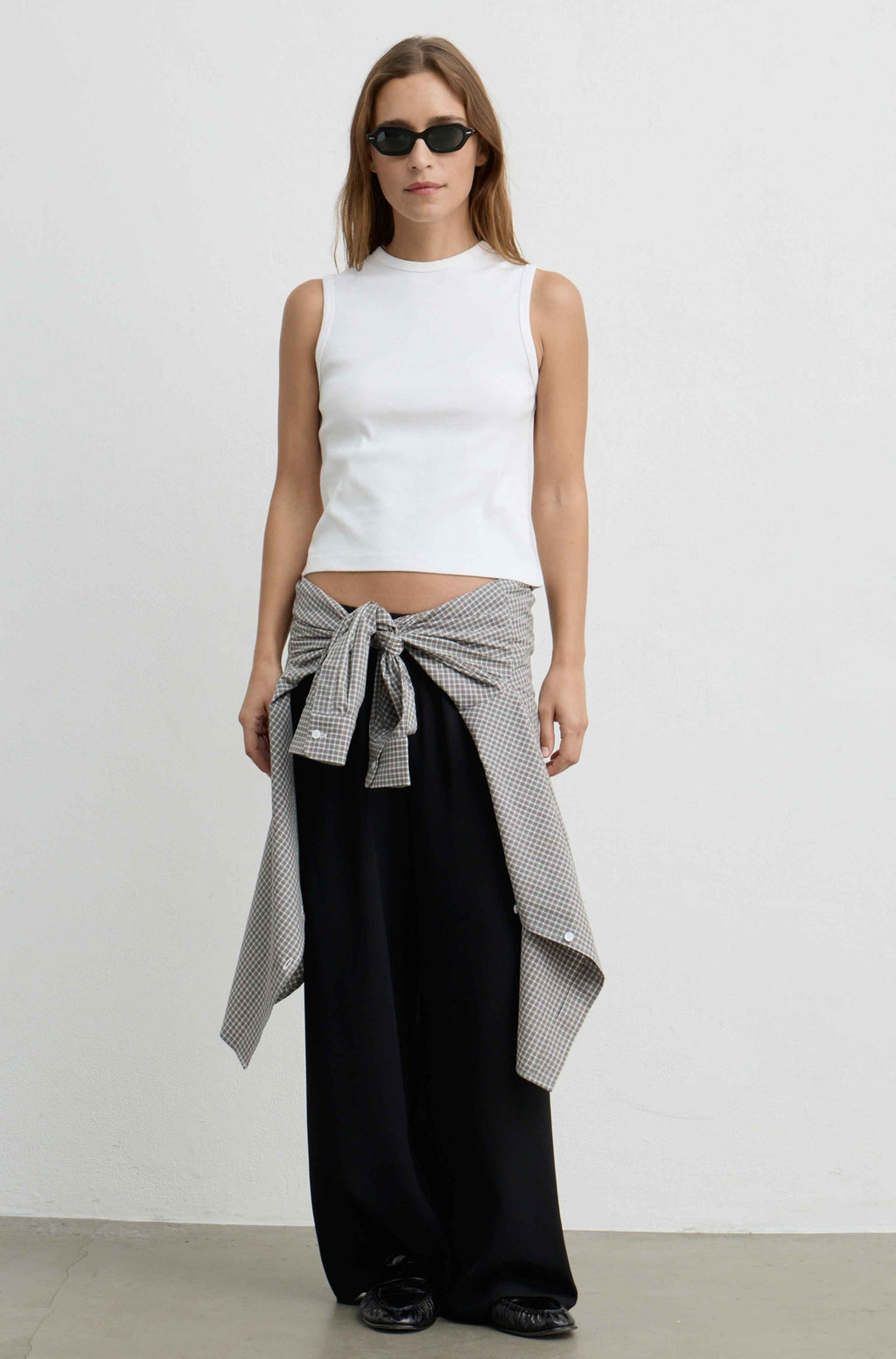 A woman stands against a plain light gray wall wearing a white sleeveless mock-neck top, loose black pants, black sunglasses, and a gray-and-white patterned shirt tied around her waist.