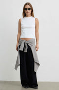 A woman stands against a plain light gray wall wearing a white sleeveless mock-neck top, loose black pants, black sunglasses, and a gray-and-white patterned shirt tied around her waist.