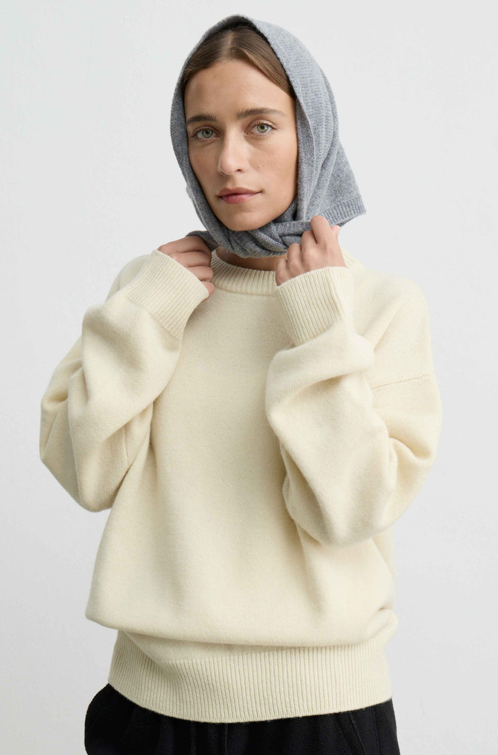 A woman wearing a yellow knit crewneck sweater and a light gray headscarf, posing against a plain light gray background.