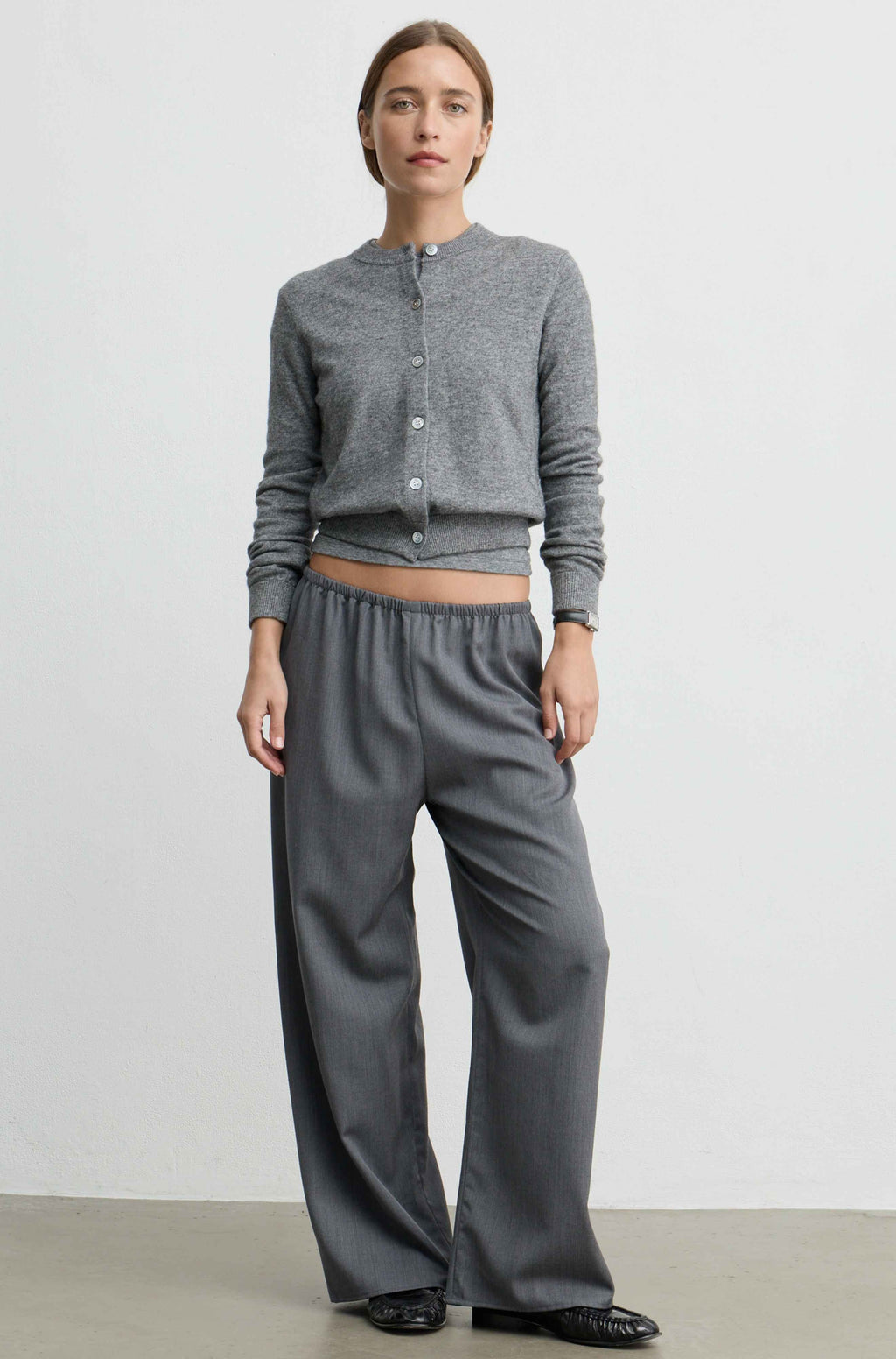 A woman modeling a gray long-sleeve cropped button-front cardigan and matching wide-leg trousers against a plain light gray studio background.