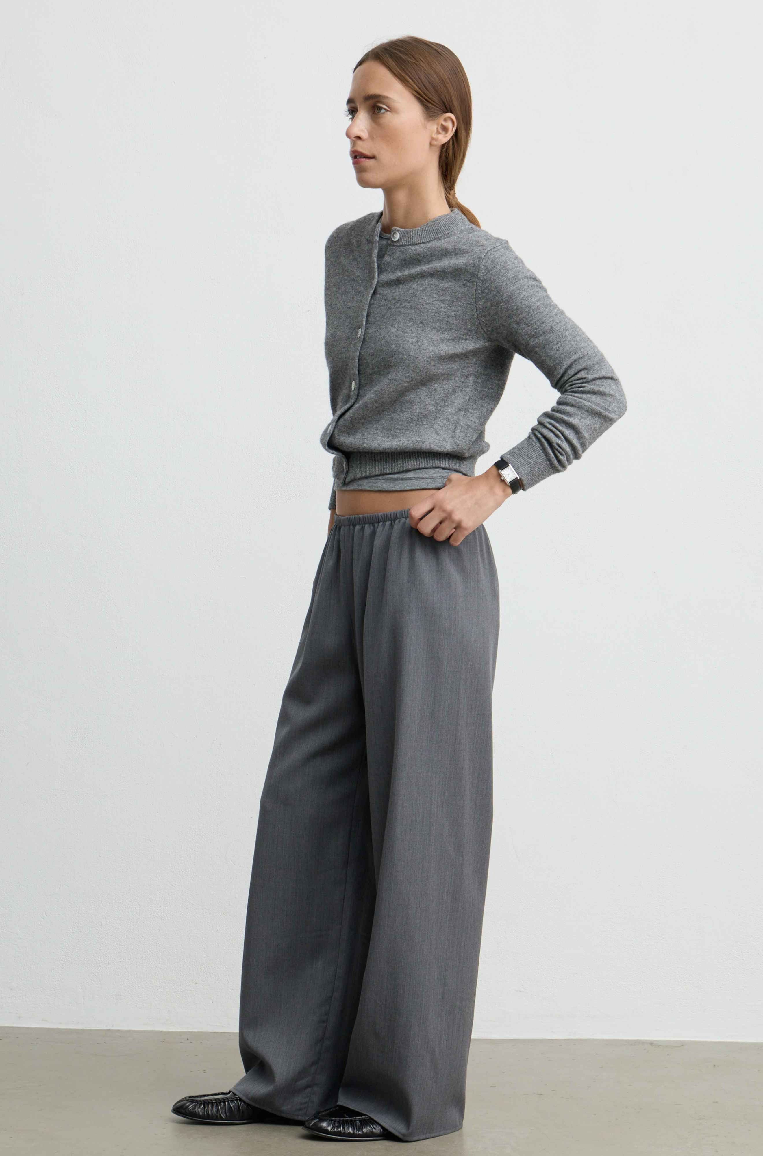 A woman modeling a gray knit long-sleeve top and matching wide-leg trousers, standing in profile against a plain light gray studio background.