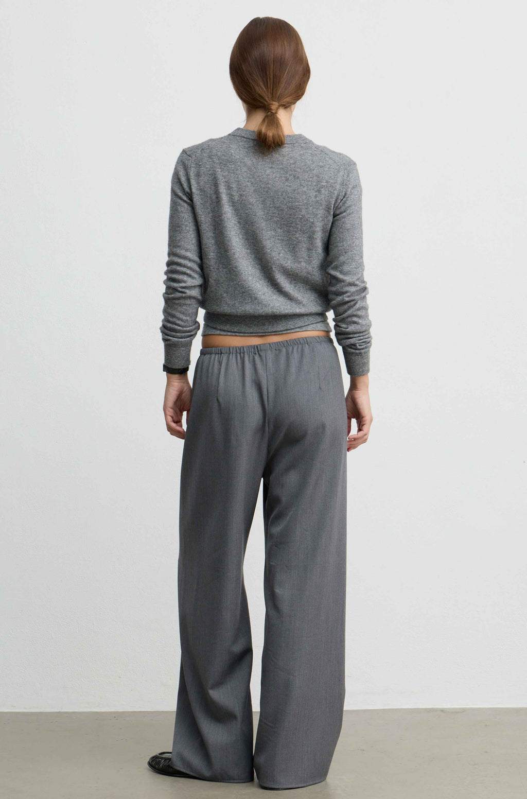 A model viewed from the back wearing a gray long-sleeve cropped sweater and matching wide-leg trousers against a plain light gray studio background.