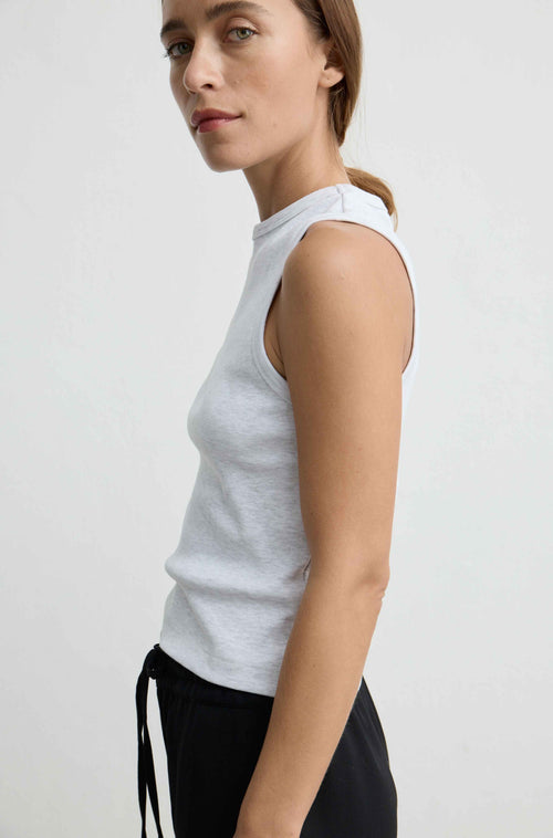 A woman shown in side profile wearing a light gray sleeveless ribbed tank top and black pants against a plain light gray studio background.