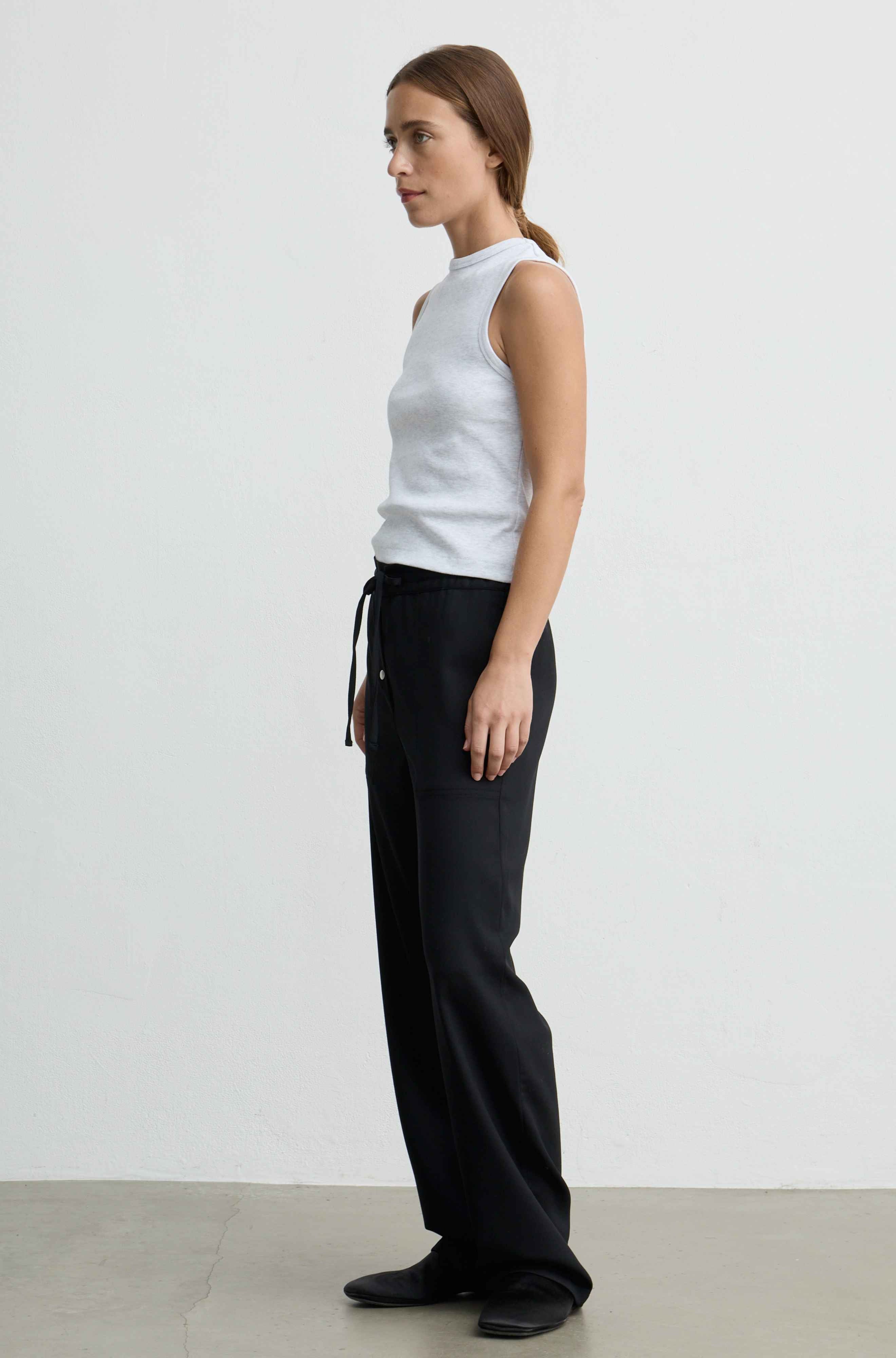 A woman stands in profile wearing a white sleeveless top and black wide-leg pants against a plain light gray studio background.