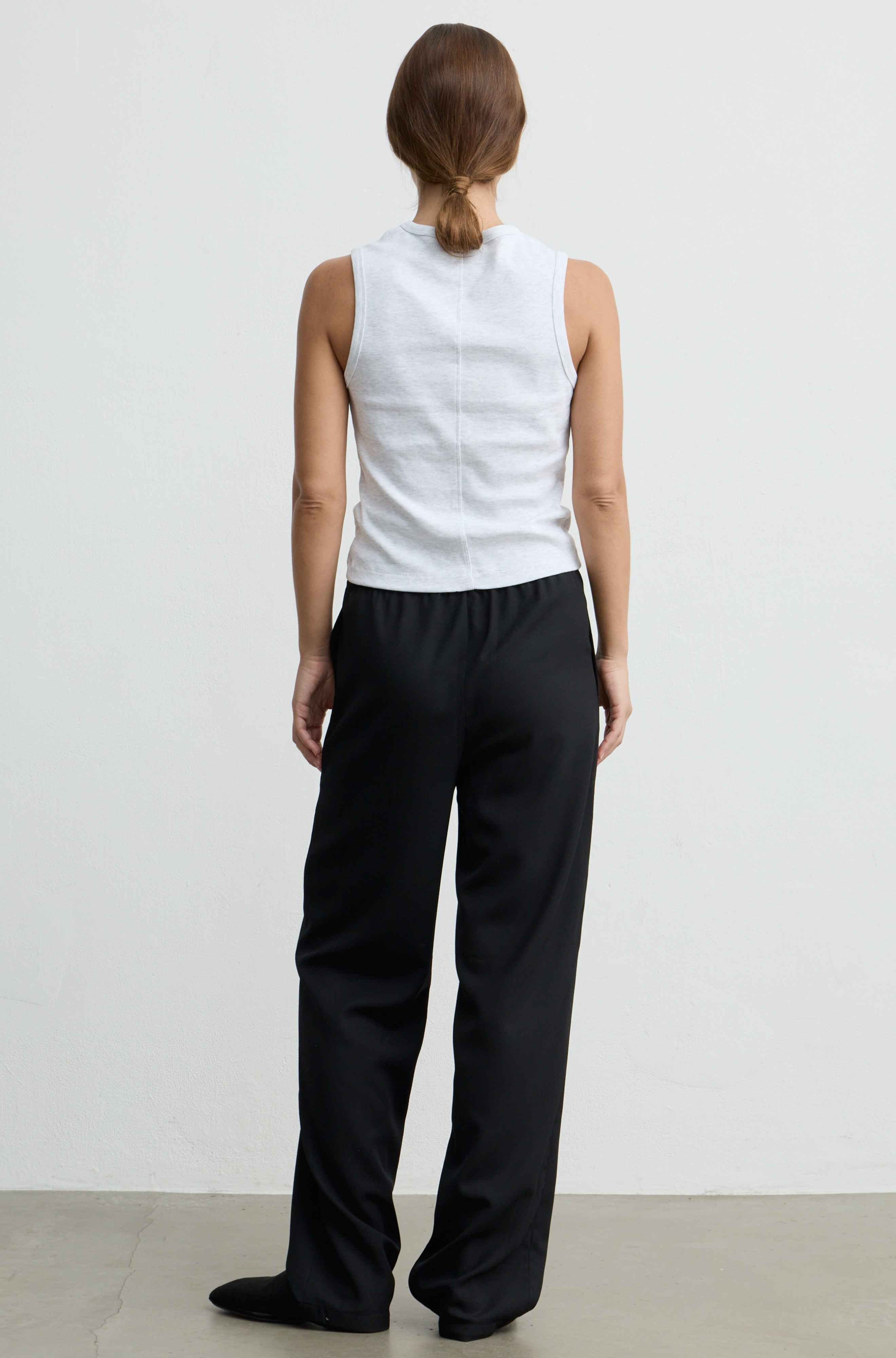 A woman shown from the back wearing a white sleeveless top and black wide-leg trousers, standing against a plain light gray wall.