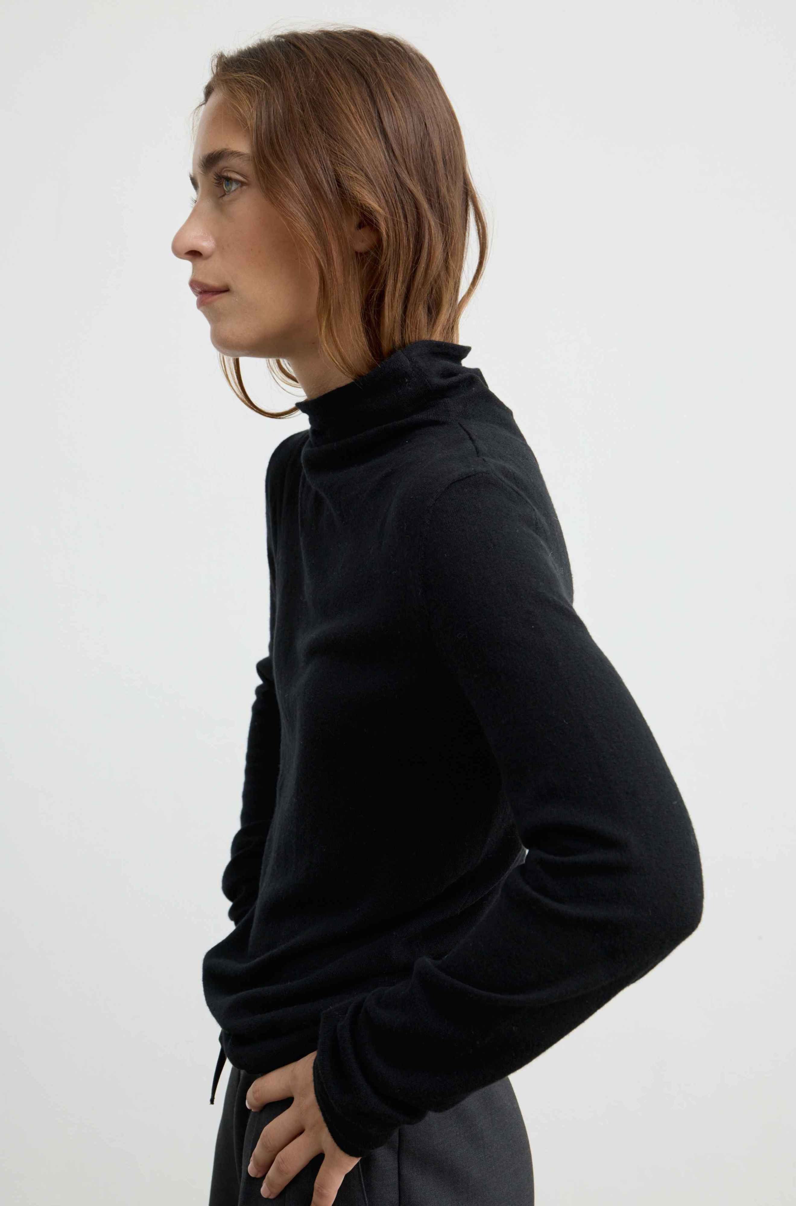 A woman in profile wearing a black long-sleeve turtleneck wool and cashmere top, photographed against a clean white studio background.