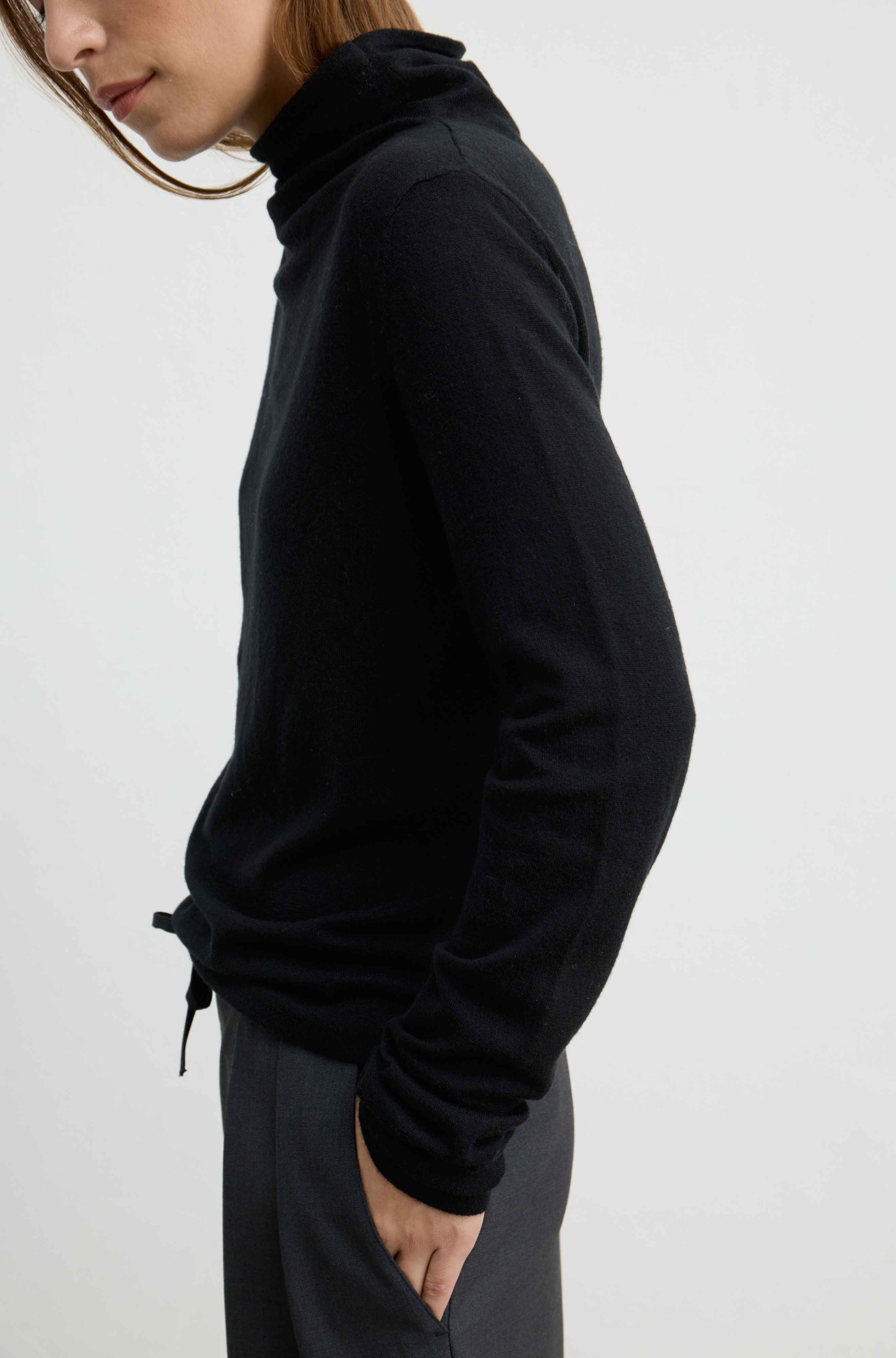 A woman shown in side profile wearing a black long-sleeve turtleneck sweater and dark gray pants against a plain light gray background.