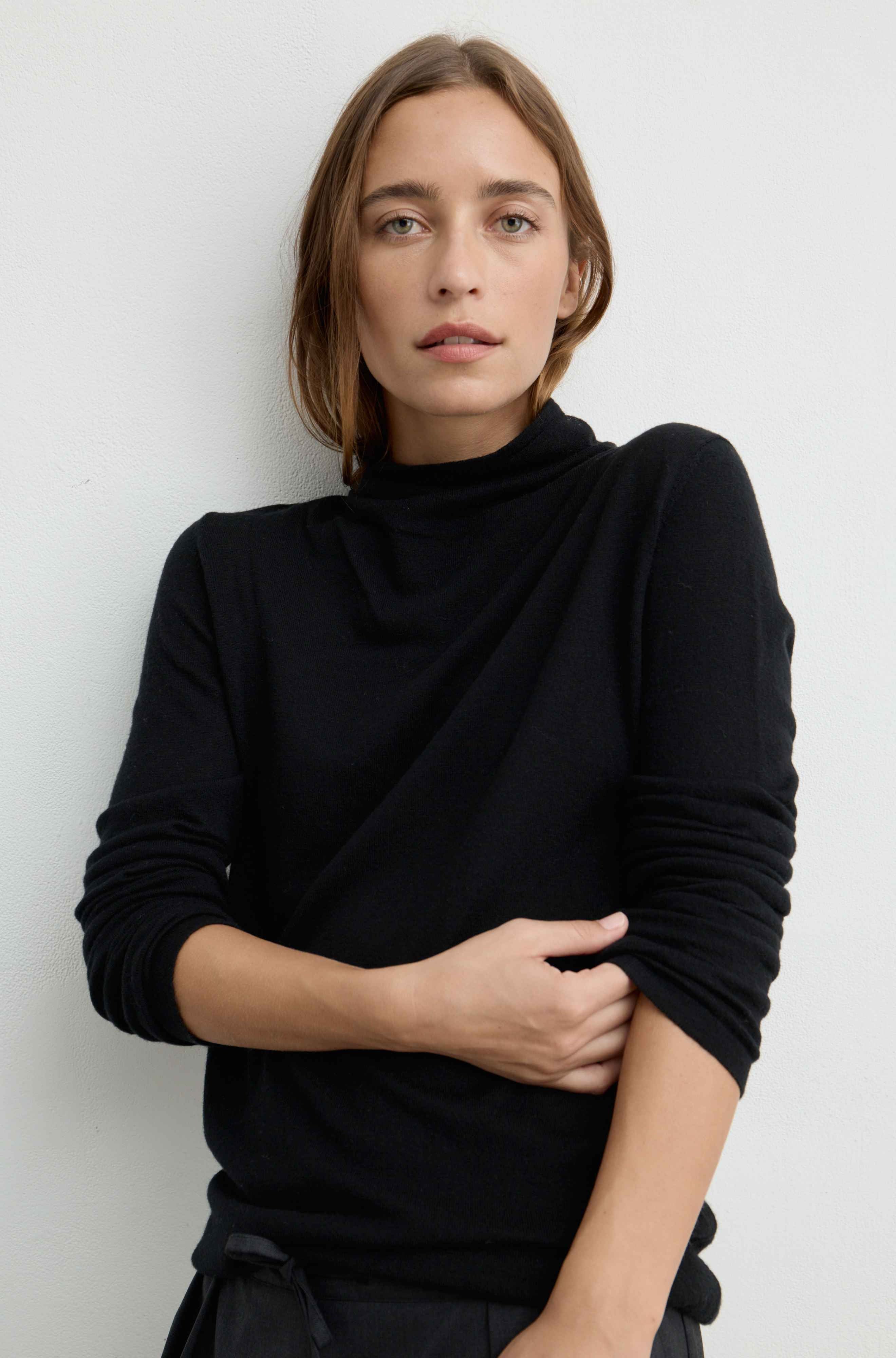 A woman with shoulder-length brown hair wearing a black long-sleeve turtleneck top, posing against a light gray wall.