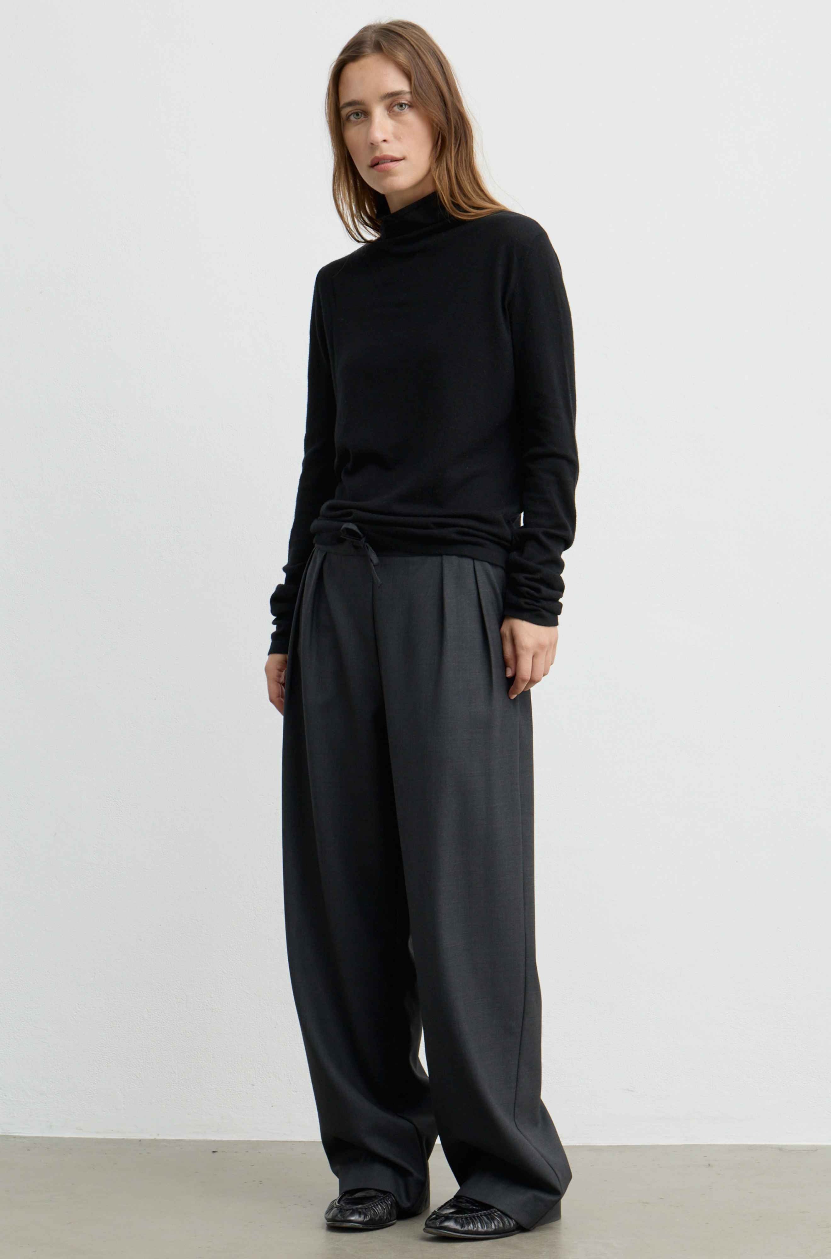 A woman modeling a black long-sleeve mock neck top and wide-leg dark gray trousers, standing against a plain light gray studio background.