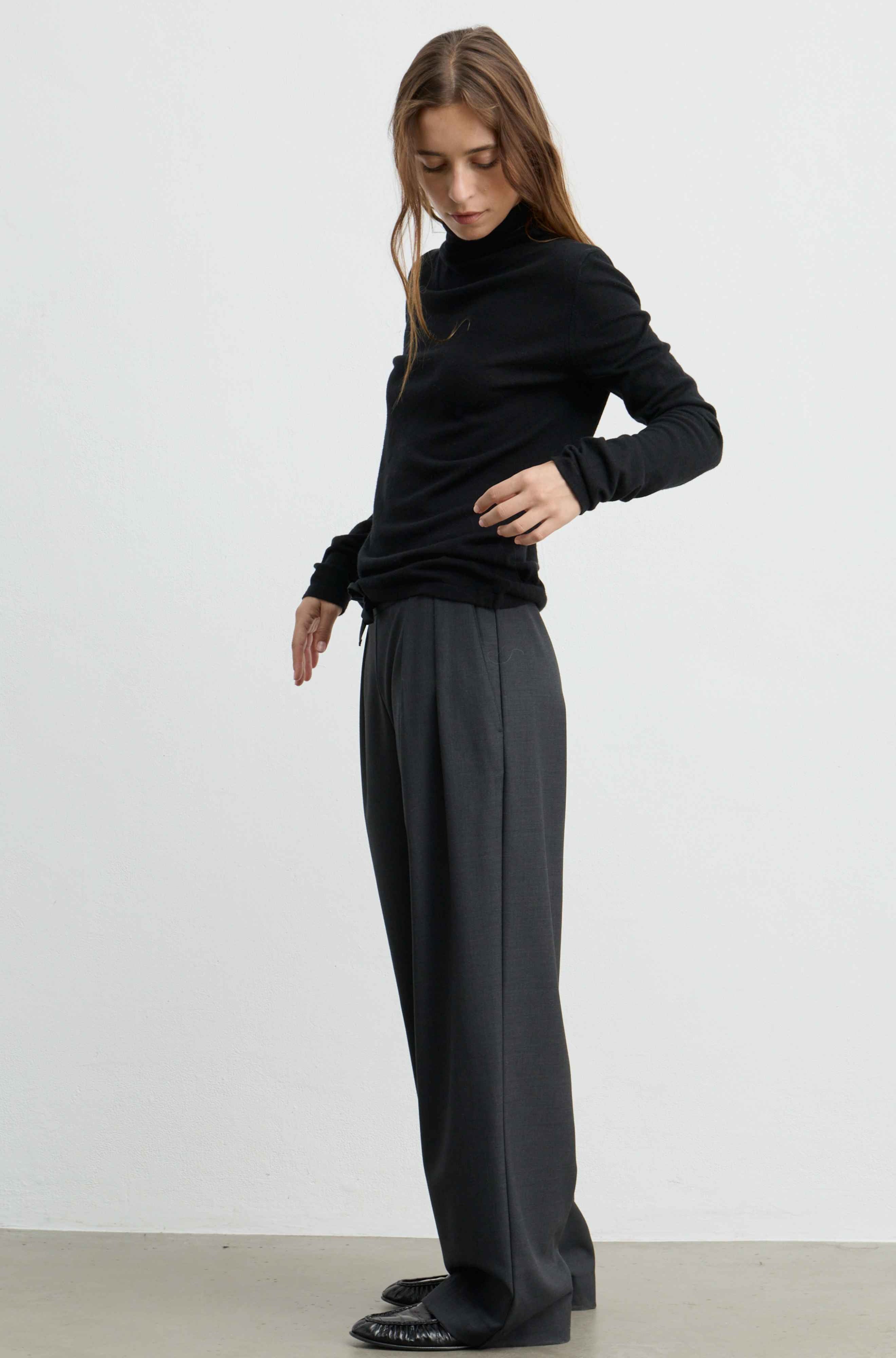 A woman modeling a black long-sleeve turtleneck sweater and dark charcoal gray wide-leg trousers, standing against a plain light gray studio background.