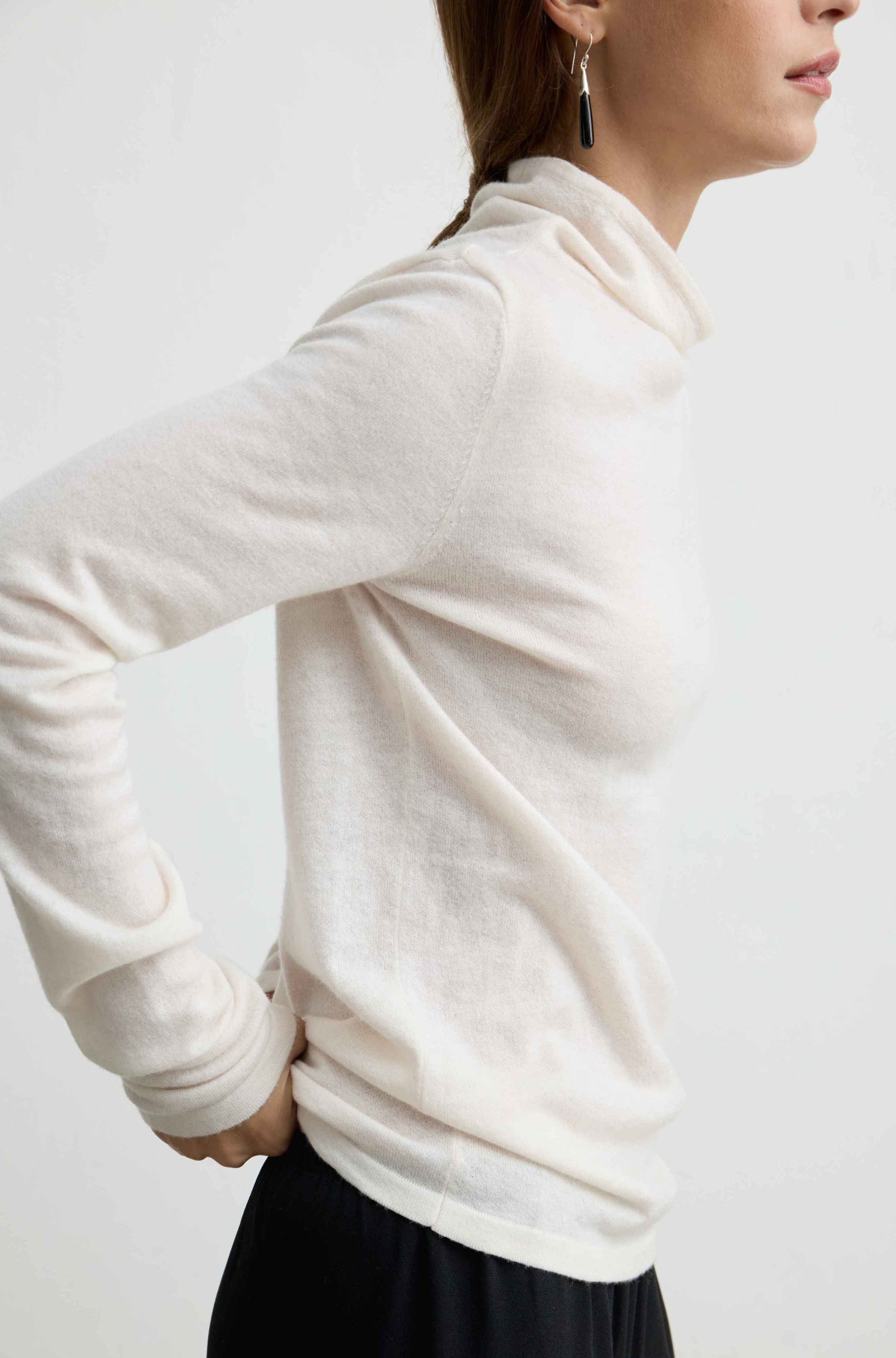 A woman wearing a cream-colored, long-sleeve turtleneck knit sweater, shown from a side-back angle against a plain white studio background.