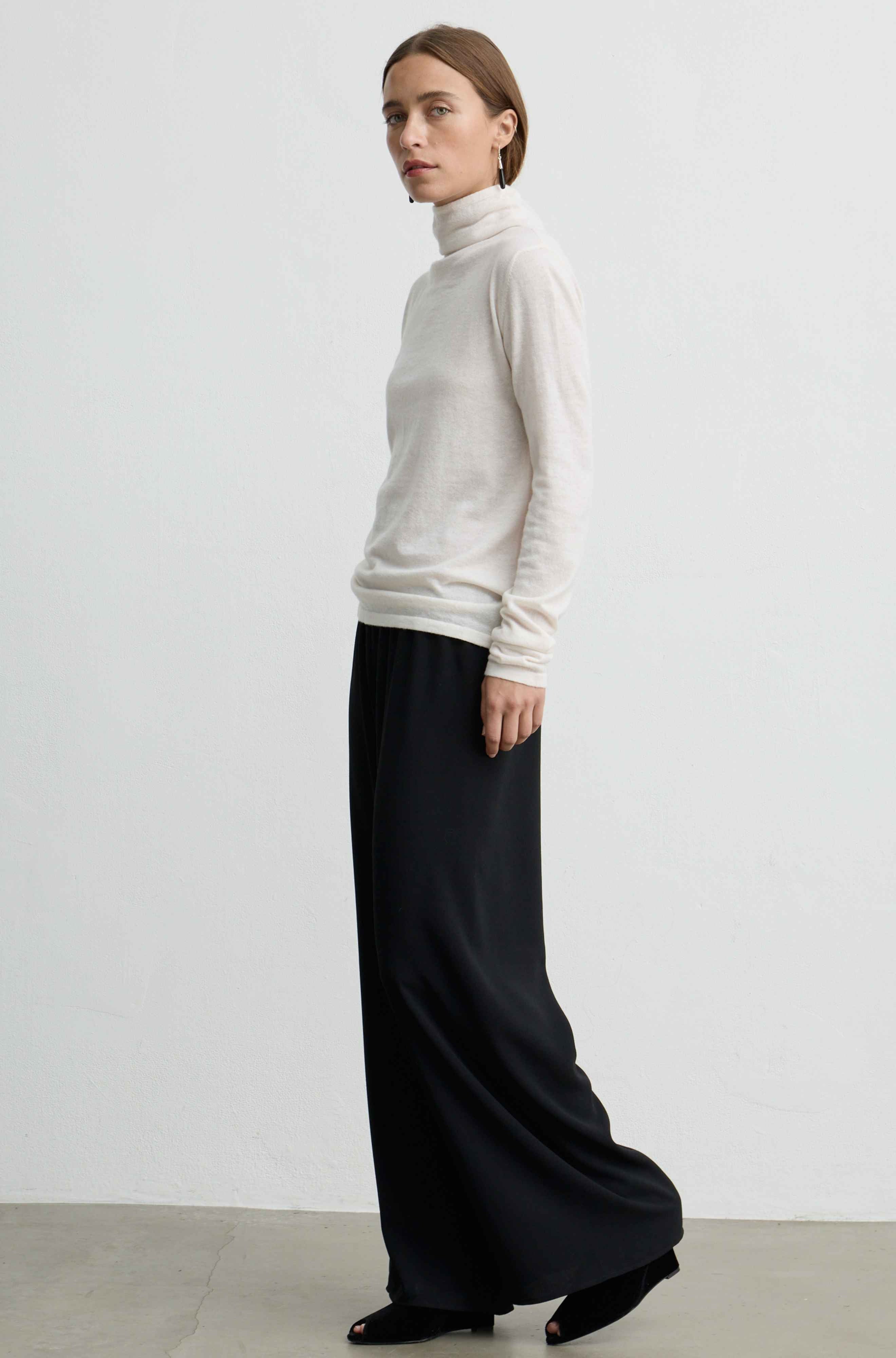 A woman stands in profile wearing a cream turtleneck sweater and wide-leg black trousers against a plain light gray studio wall.