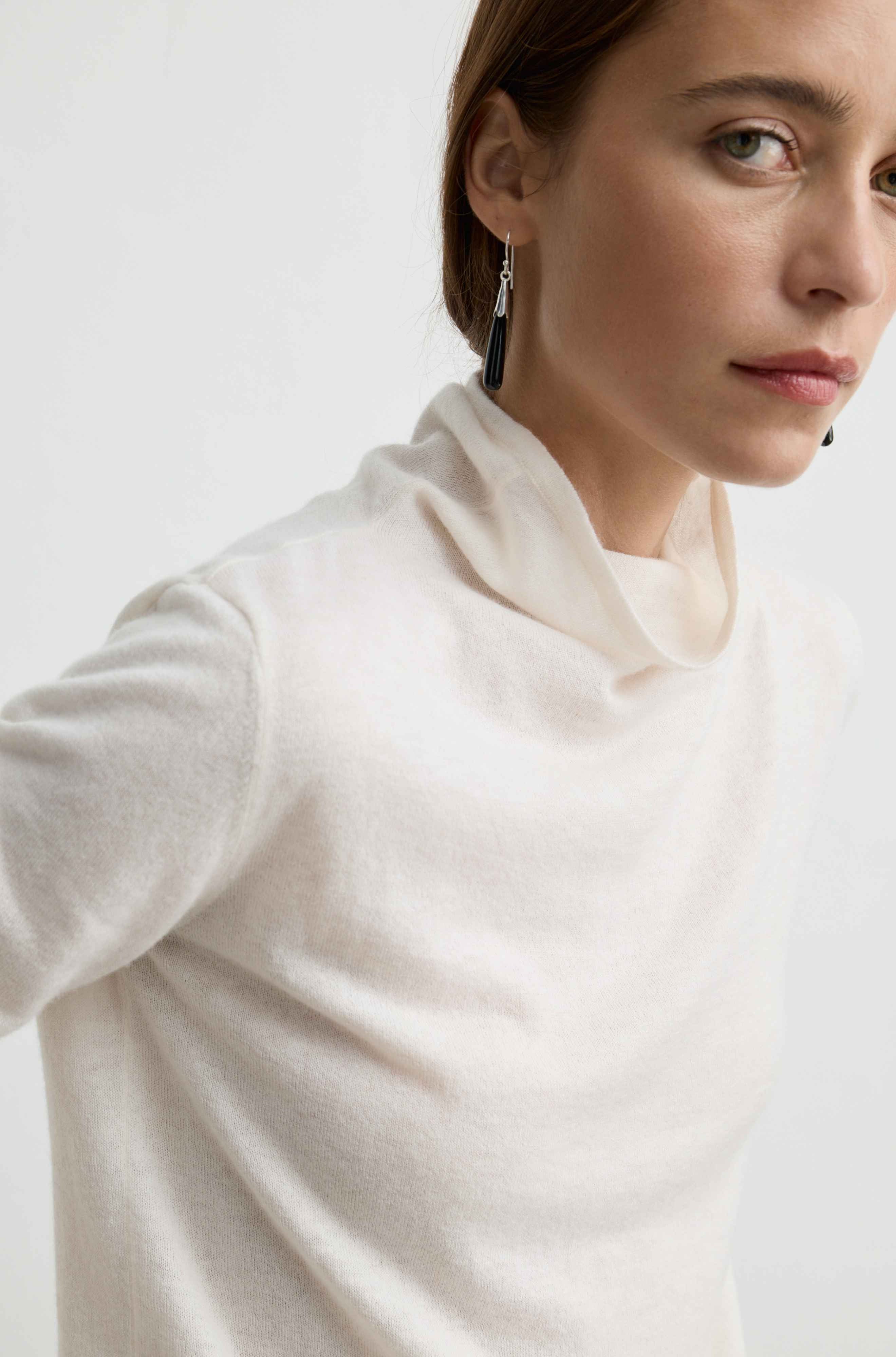 A close-up of a woman wearing a soft off-white cowl-neck knit top against a plain light background.