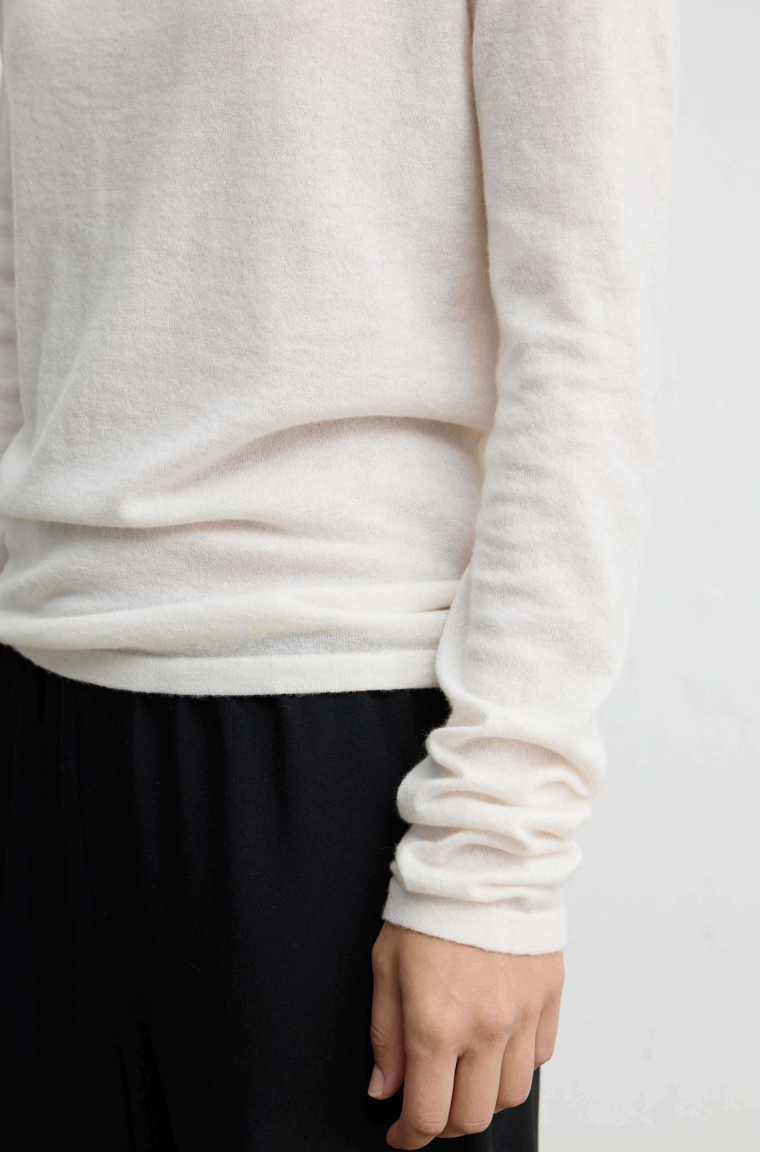 Close-up of a person wearing a soft off-white long-sleeve knit top with gathered cuffs, paired with dark navy pants, against a plain light background.