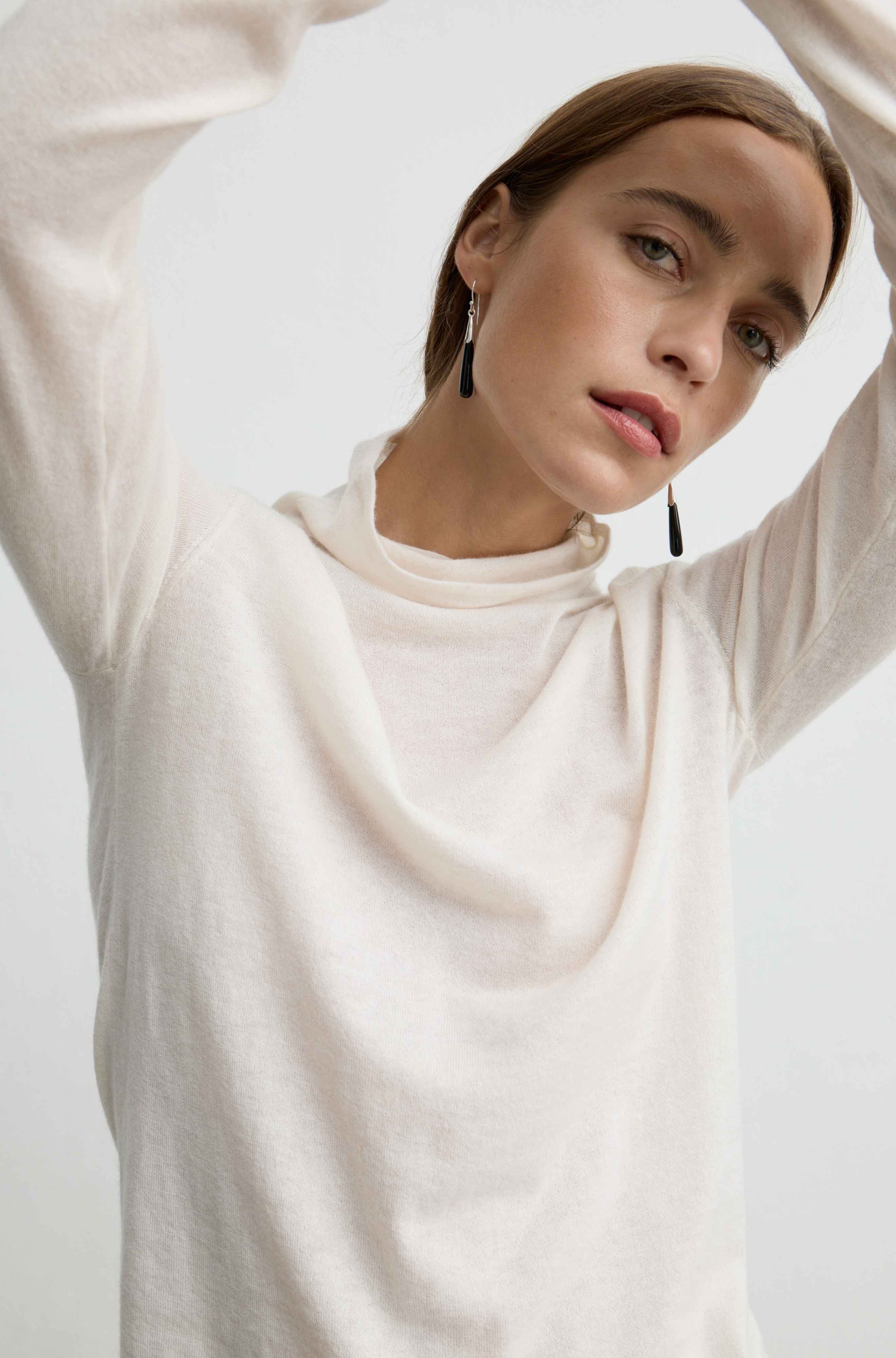 A woman wearing a cream-colored, long-sleeve cowl-neck knit top, posing with her arms raised against a plain light background.