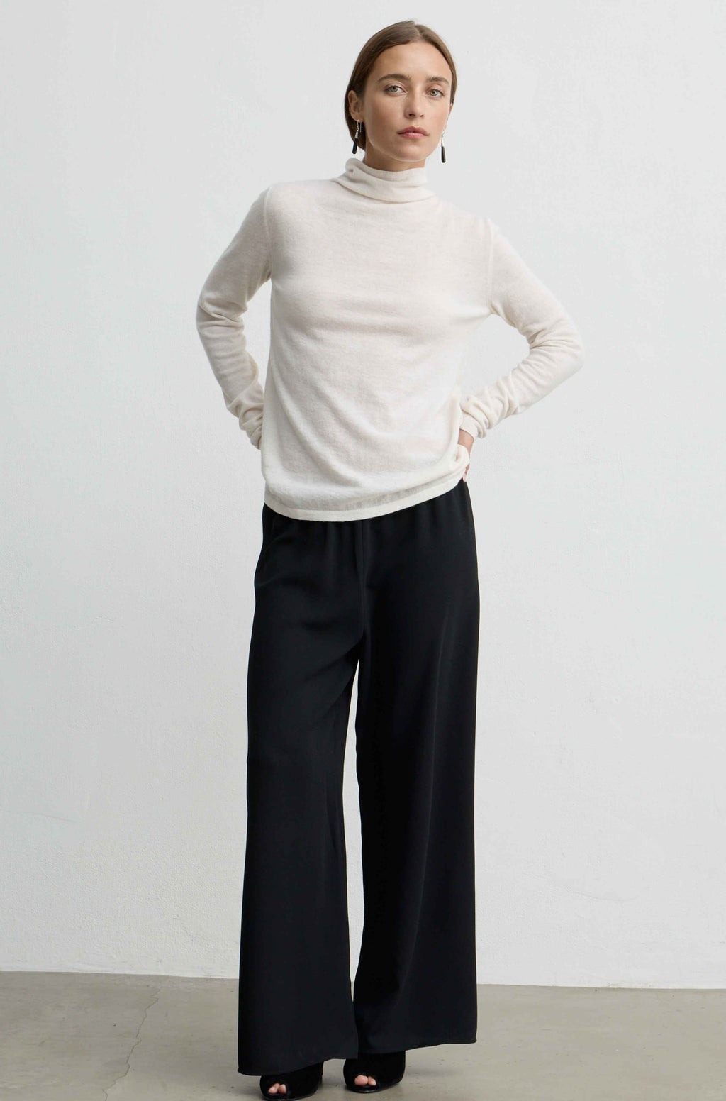 A woman modeling a white long-sleeve turtleneck sweater paired with black wide-leg trousers, standing against a plain light gray studio wall.