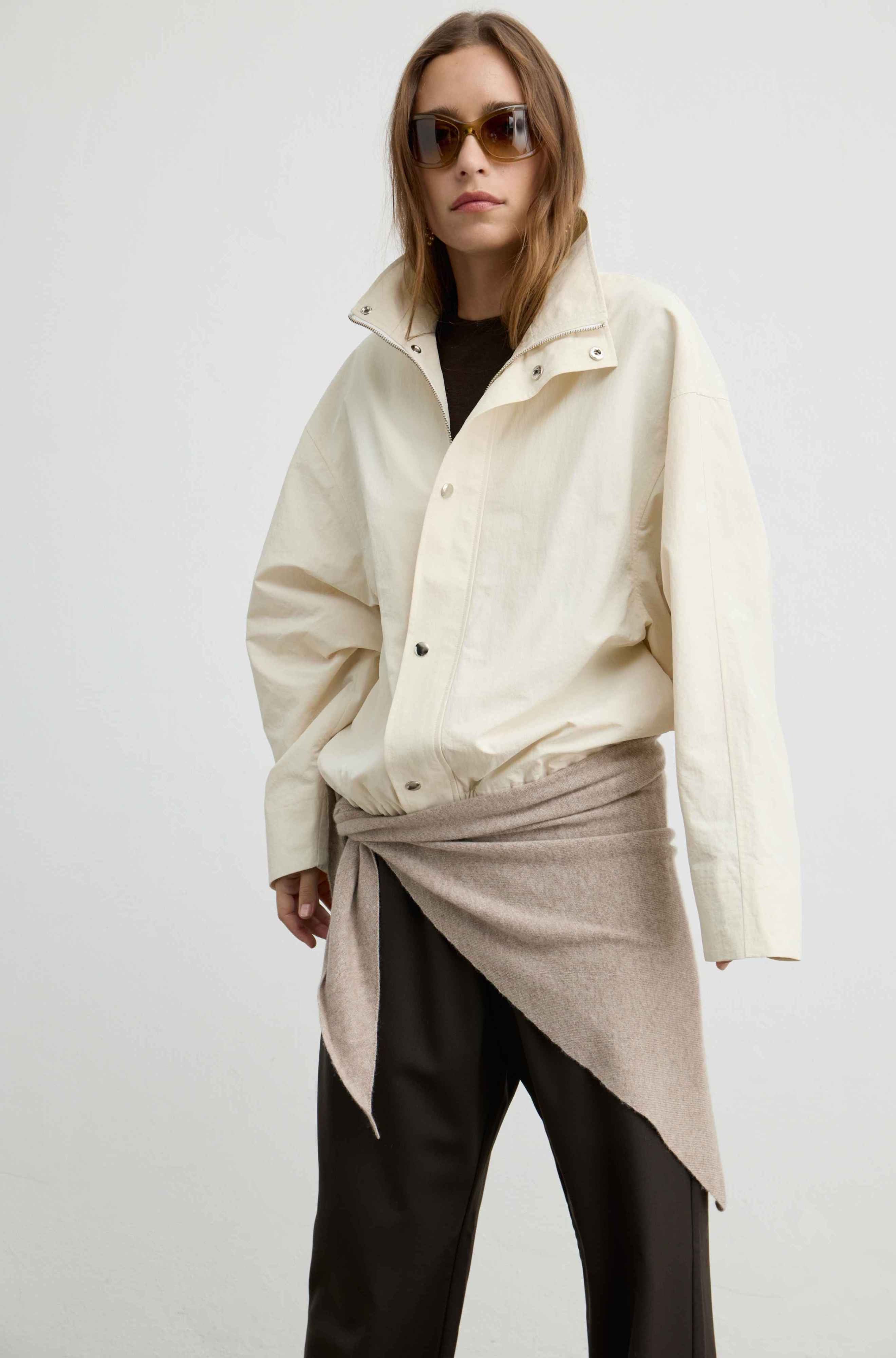 A model wearing an oversized cream-colored utility jacket with a high collar, styled with a beige wrap tied around the waist, dark trousers, and sunglasses against a plain light gray background.