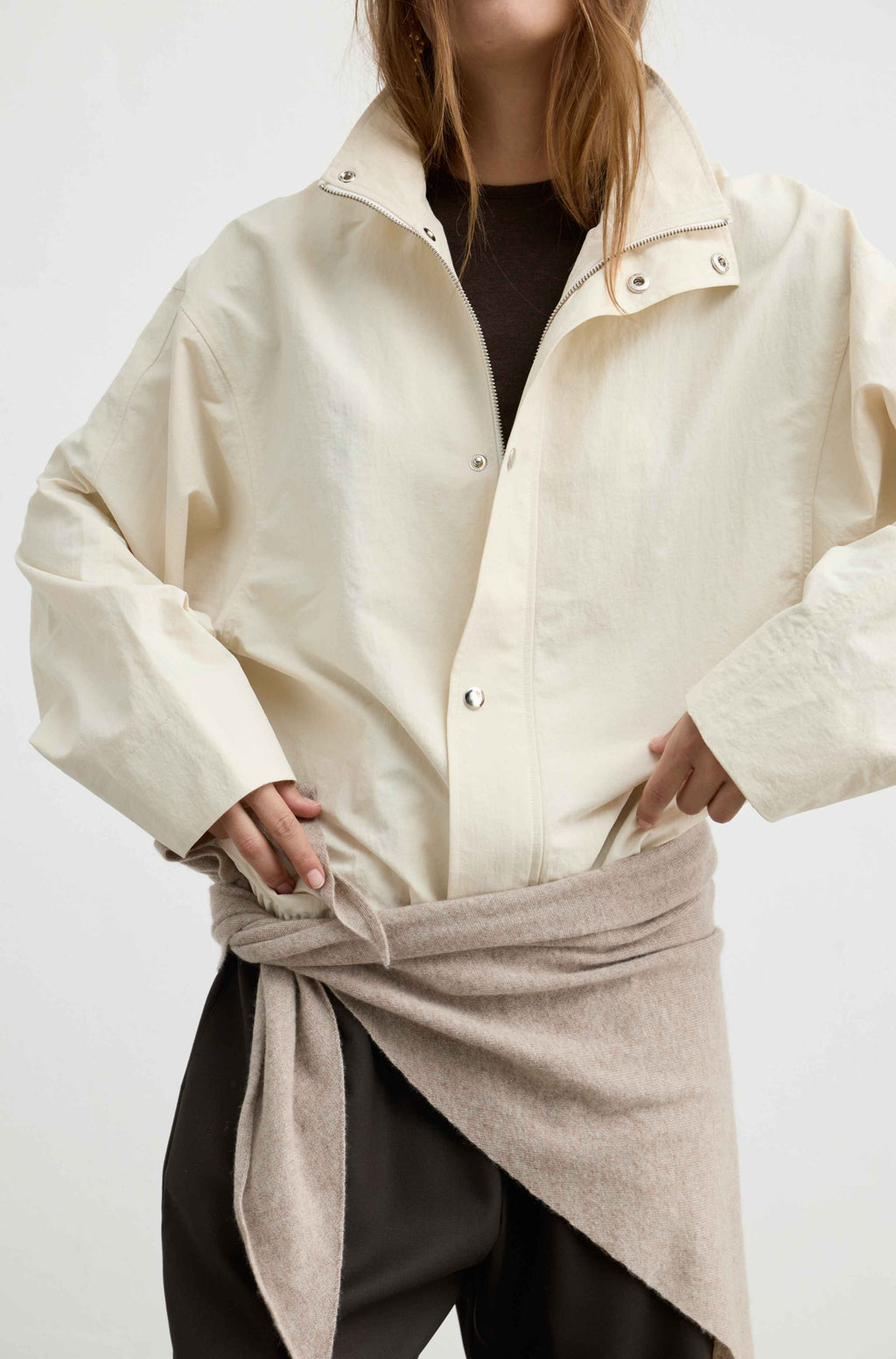 A model wearing a cream-colored oversized button-up shirt jacket with a high collar, styled with a beige wrap tied around the waist over a black top and black pants.