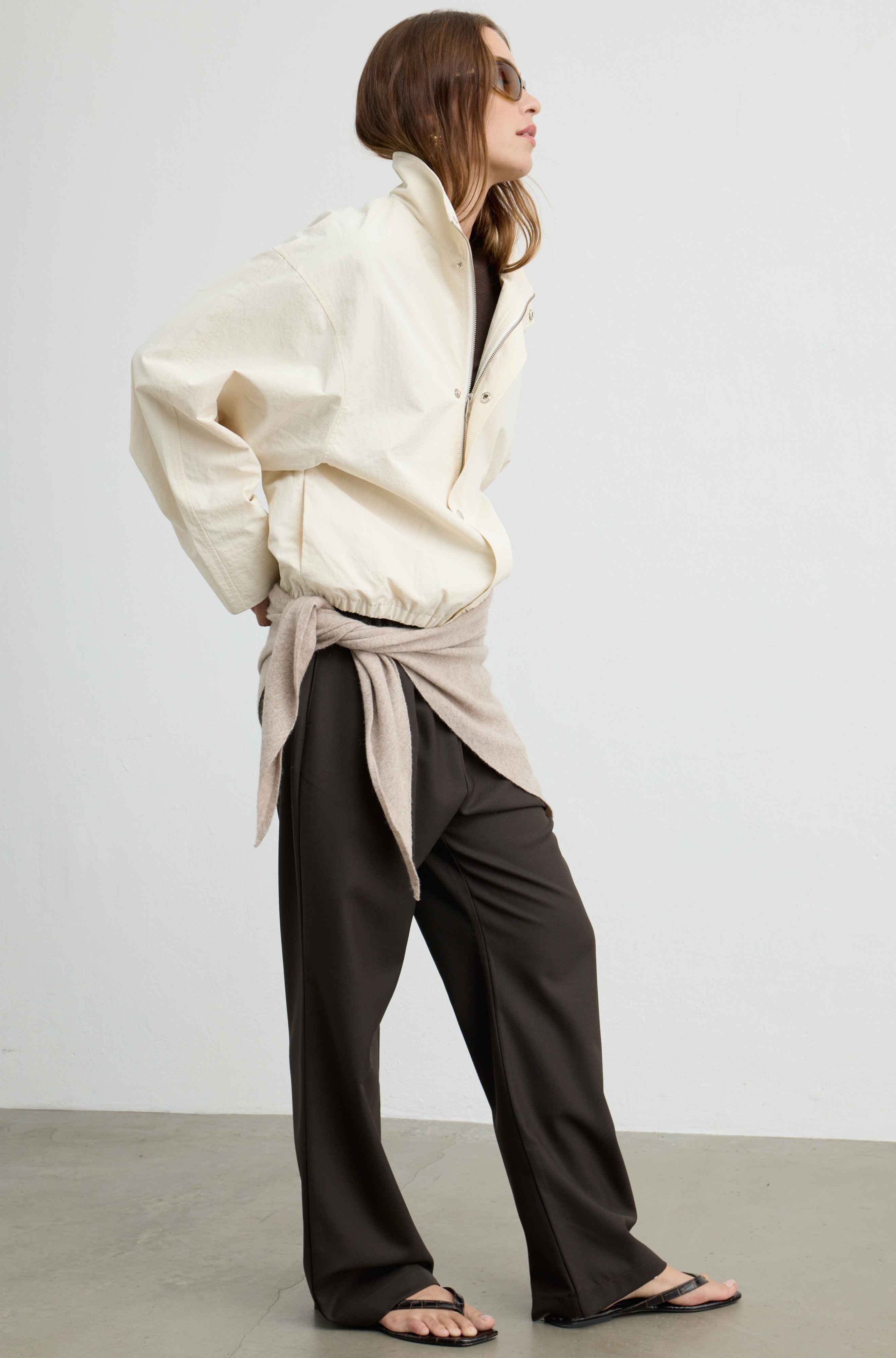 A model is shown from the side wearing a cream-colored oversized zip-up jacket, dark brown wide-leg trousers, and black sandals, with a beige shirt tied around her waist, set against a minimalist studio background.