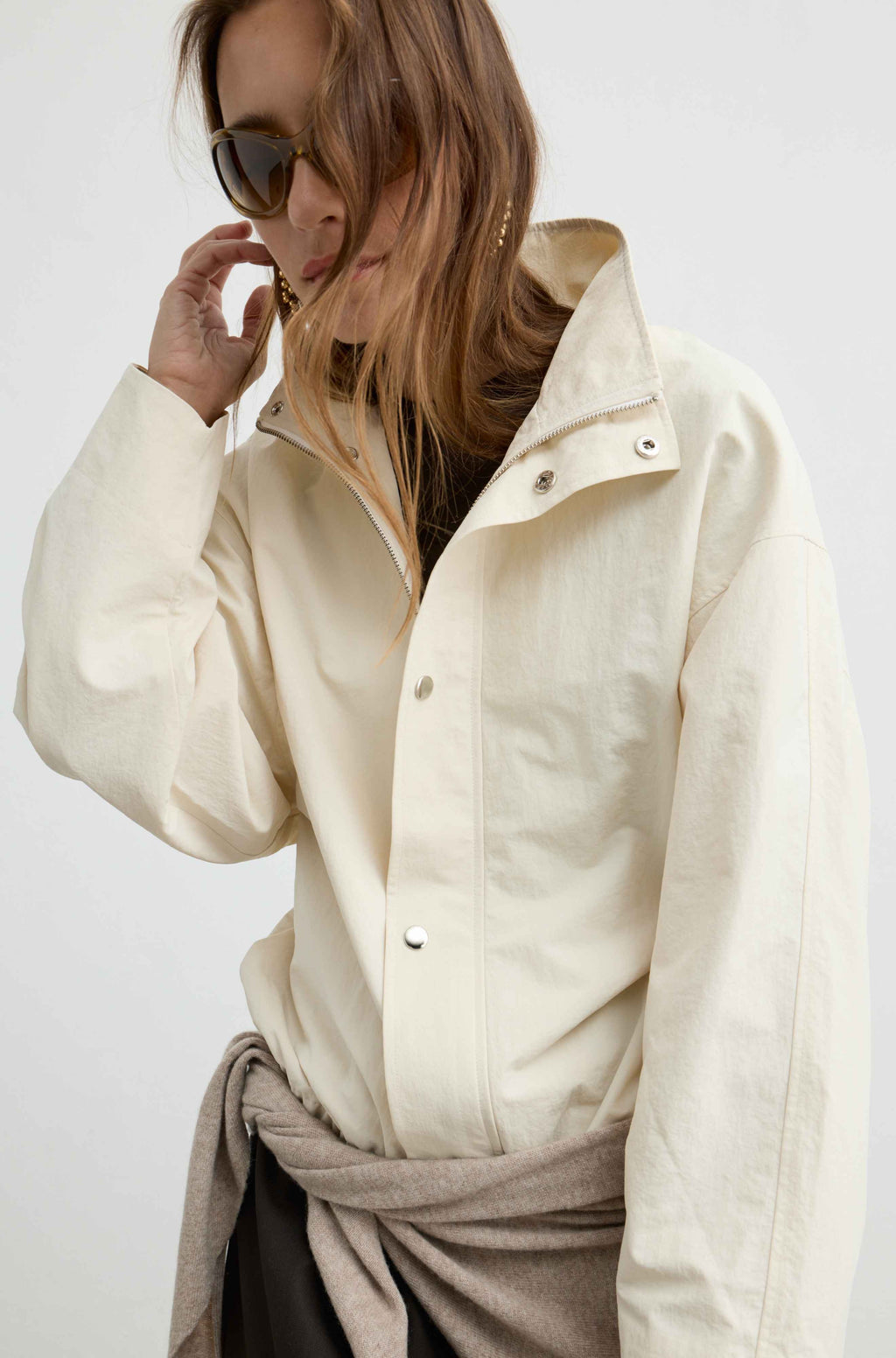A woman wearing an oversized cream-colored linen button-up shirt with a high collar and silver snap buttons, styled with a taupe scarf tied around her waist and dark sunglasses.