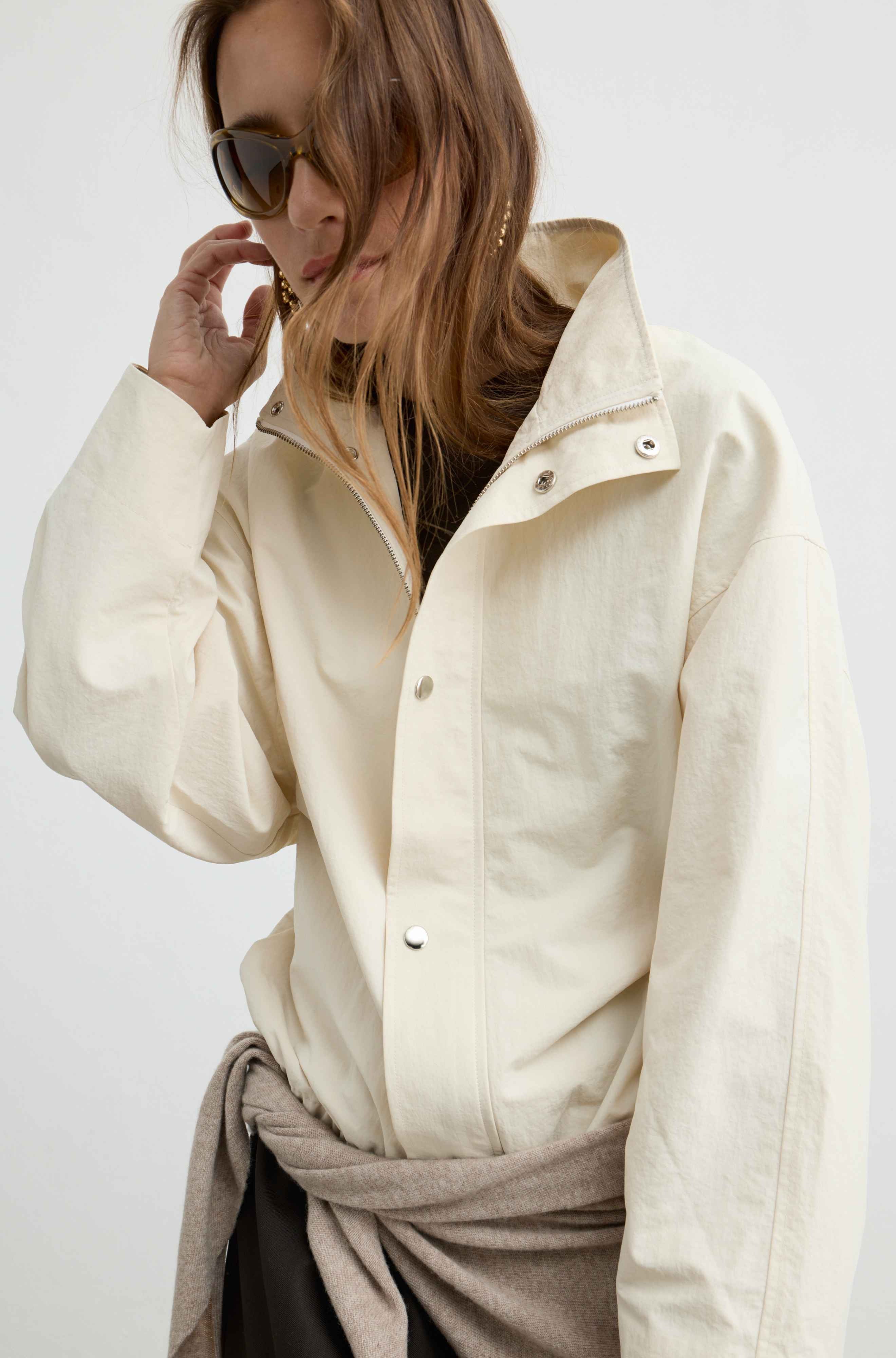 A woman wearing an oversized cream-colored linen button-up shirt with a high collar and silver snap buttons, styled with a taupe scarf tied around her waist and dark sunglasses.