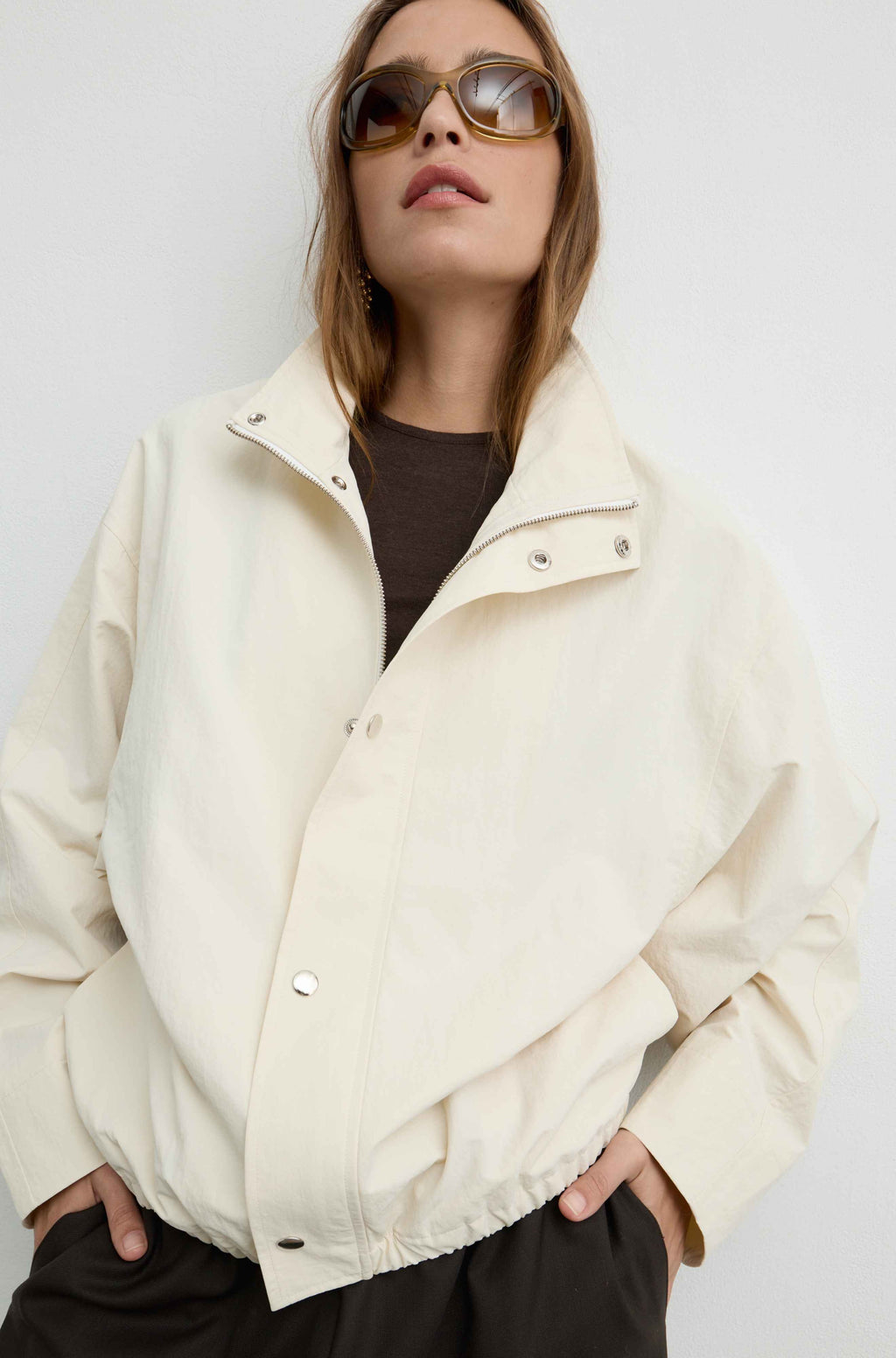 A woman wearing a cream-colored lightweight jacket over a dark brown t-shirt and brown pants, styled with tortoiseshell sunglasses, against a plain light gray background.