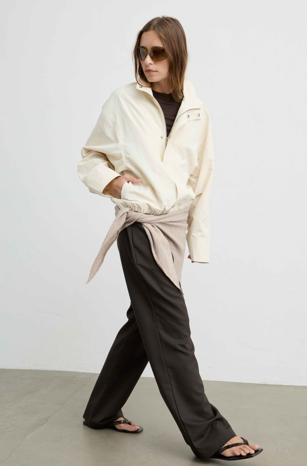 A woman modeling a cream zip-up overshirt tied at the waist over a black top and dark wide-leg trousers, photographed in a minimalist studio setting.