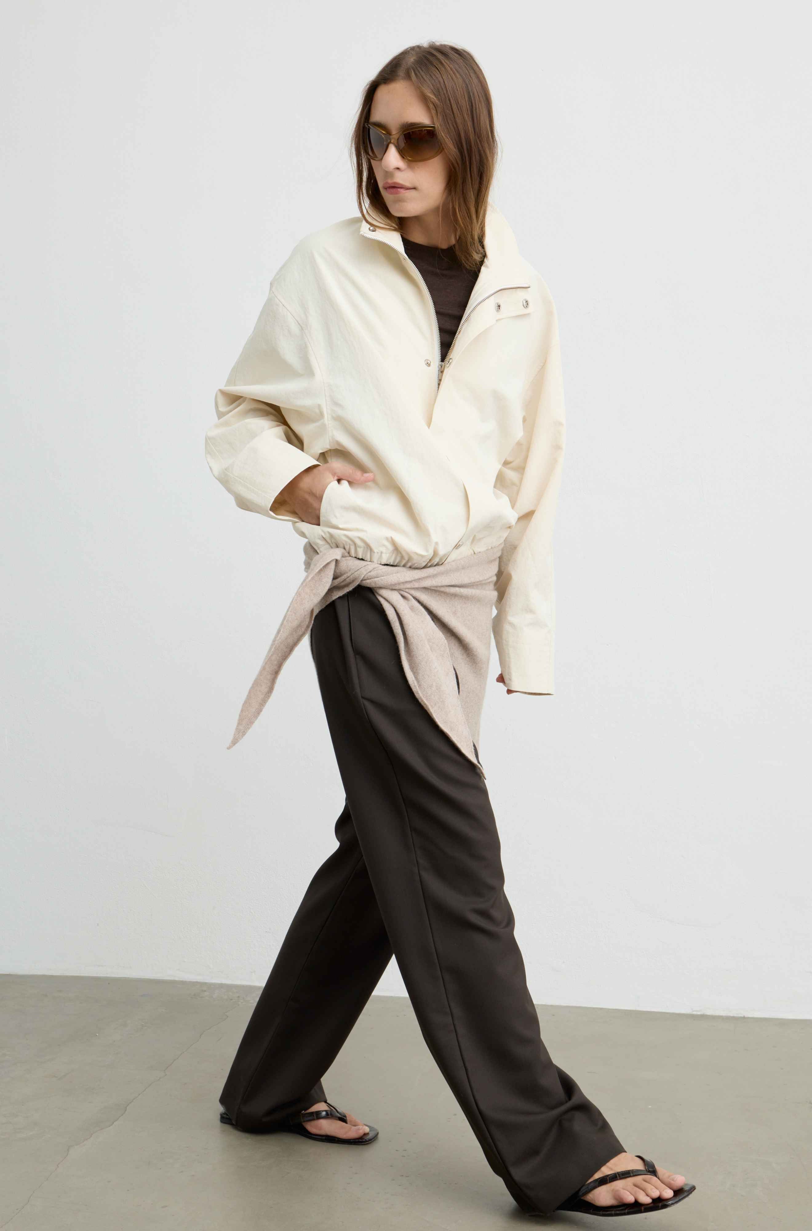 A woman modeling a cream zip-up overshirt tied at the waist over a black top and dark wide-leg trousers, photographed in a minimalist studio setting.