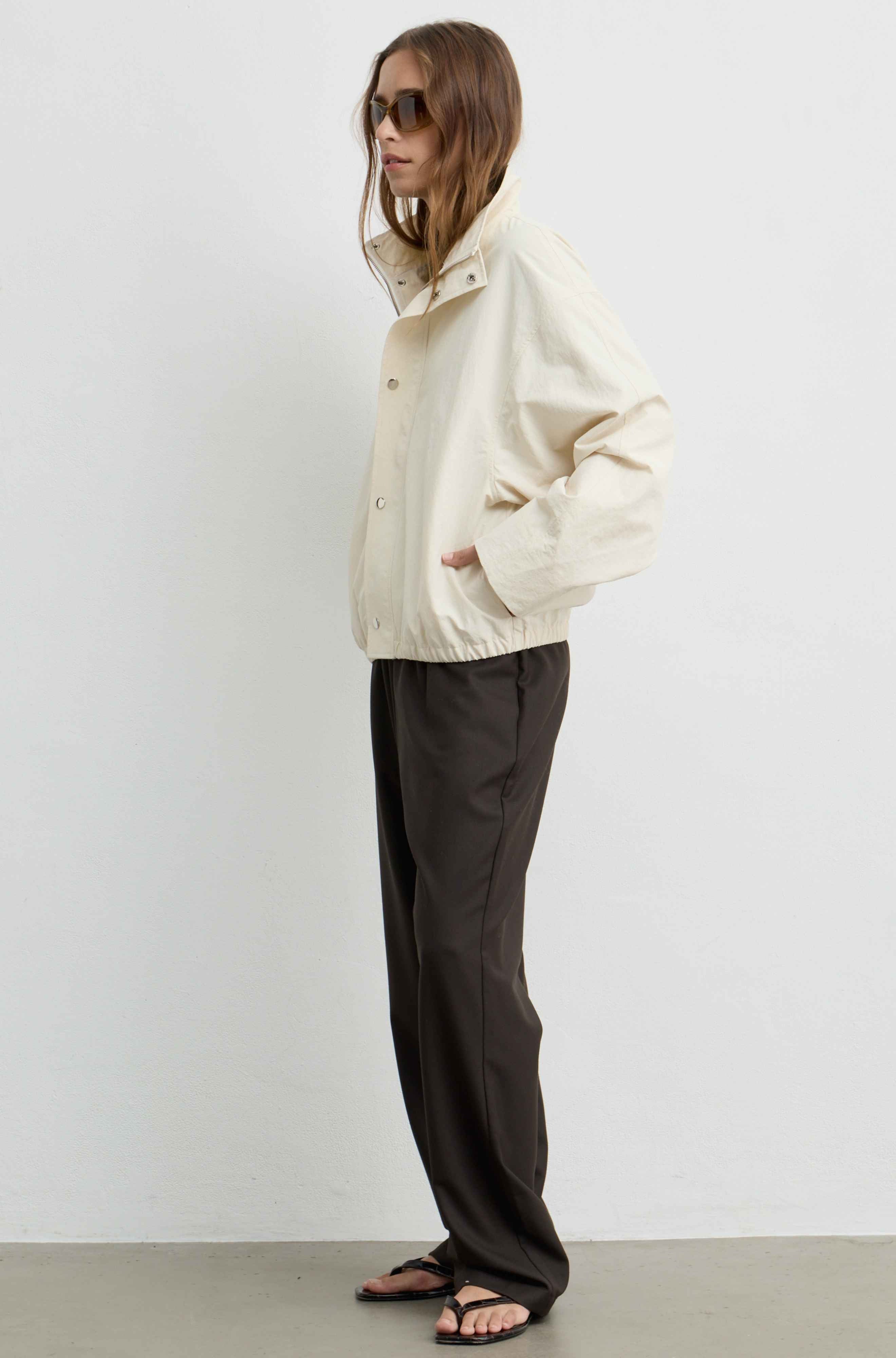 A model wearing a cream-colored utility jacket, dark brown wide-leg trousers, and black sandals, standing in profile against a plain white wall.