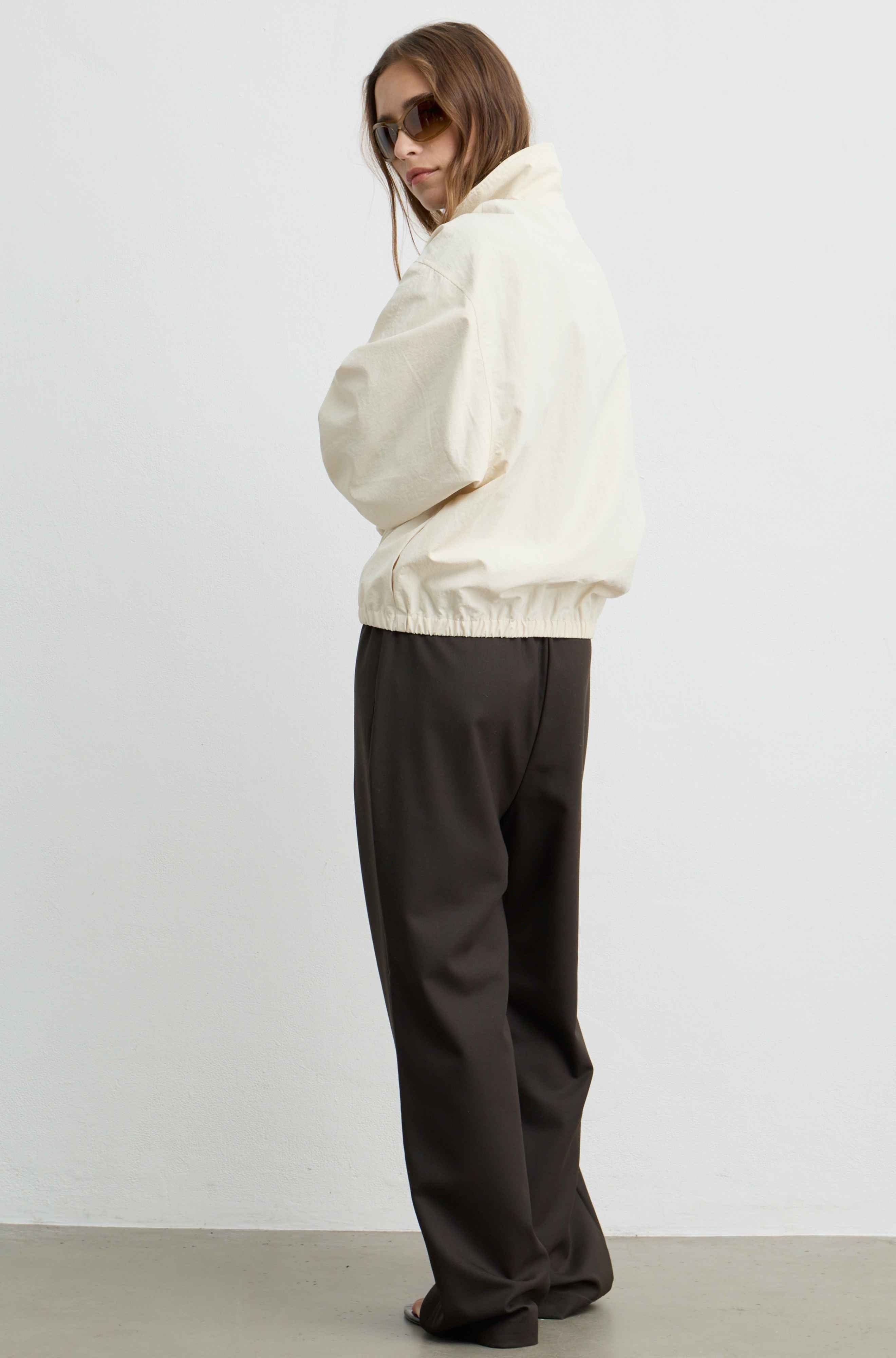 A model wearing a cream-colored windbreaker jacket and dark brown wide-leg trousers, standing in profile against a plain white studio wall.