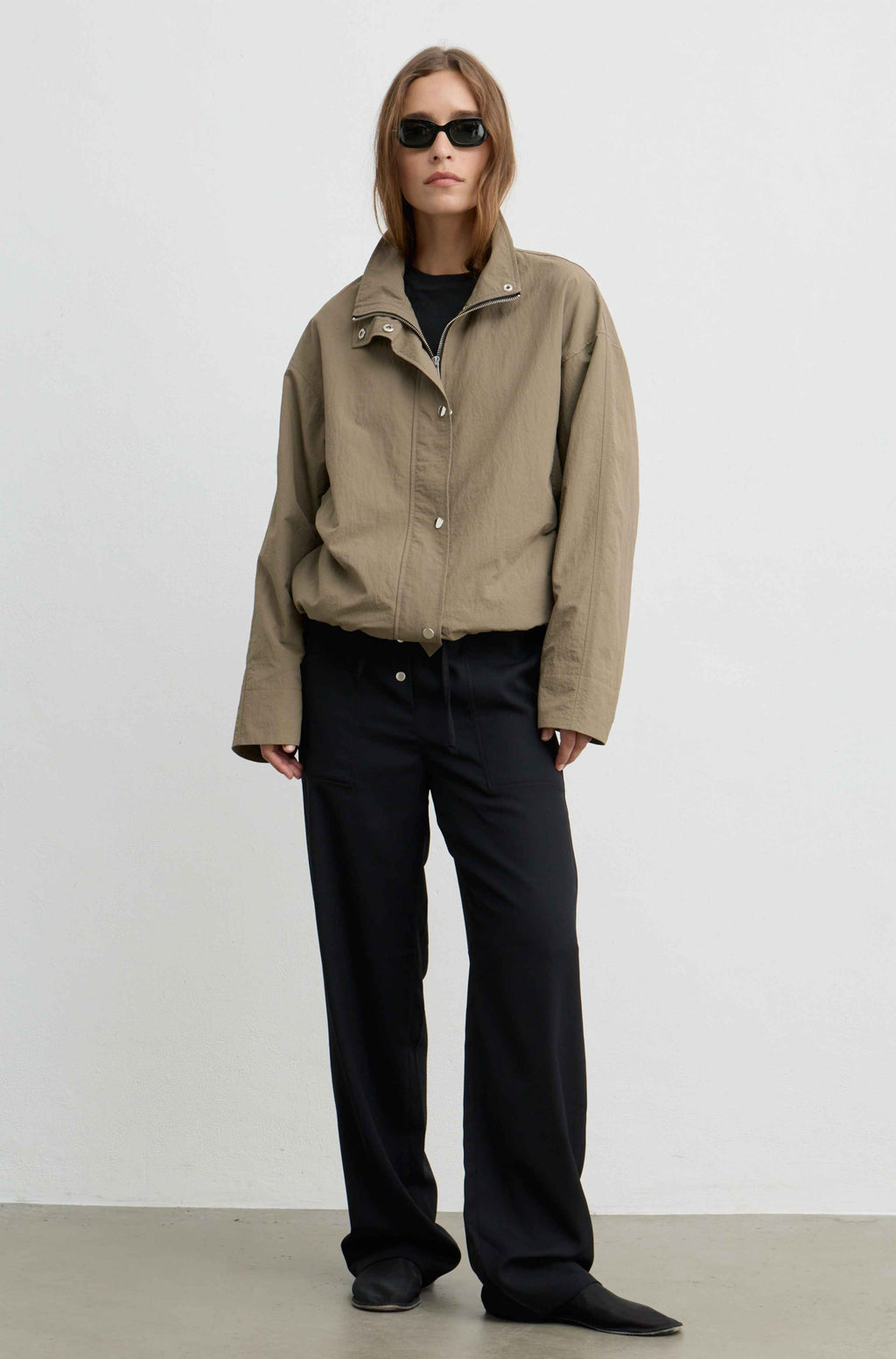 A model stands against a plain light gray studio wall wearing an oversized beige utility jacket over a black top and black wide-leg trousers.