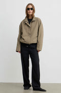 A model stands against a plain light gray studio wall wearing an oversized beige utility jacket over a black top and black wide-leg trousers.