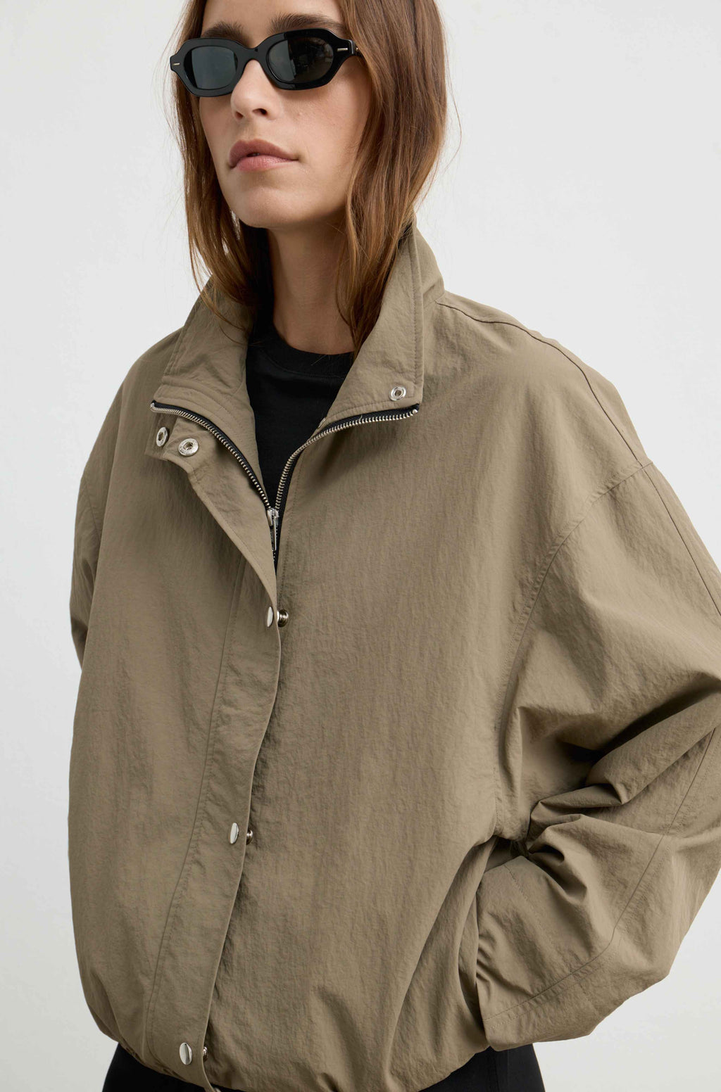A woman wearing a khaki utility jacket with a high collar and silver snap buttons over a black top, styled with black sunglasses, against a plain light gray background.