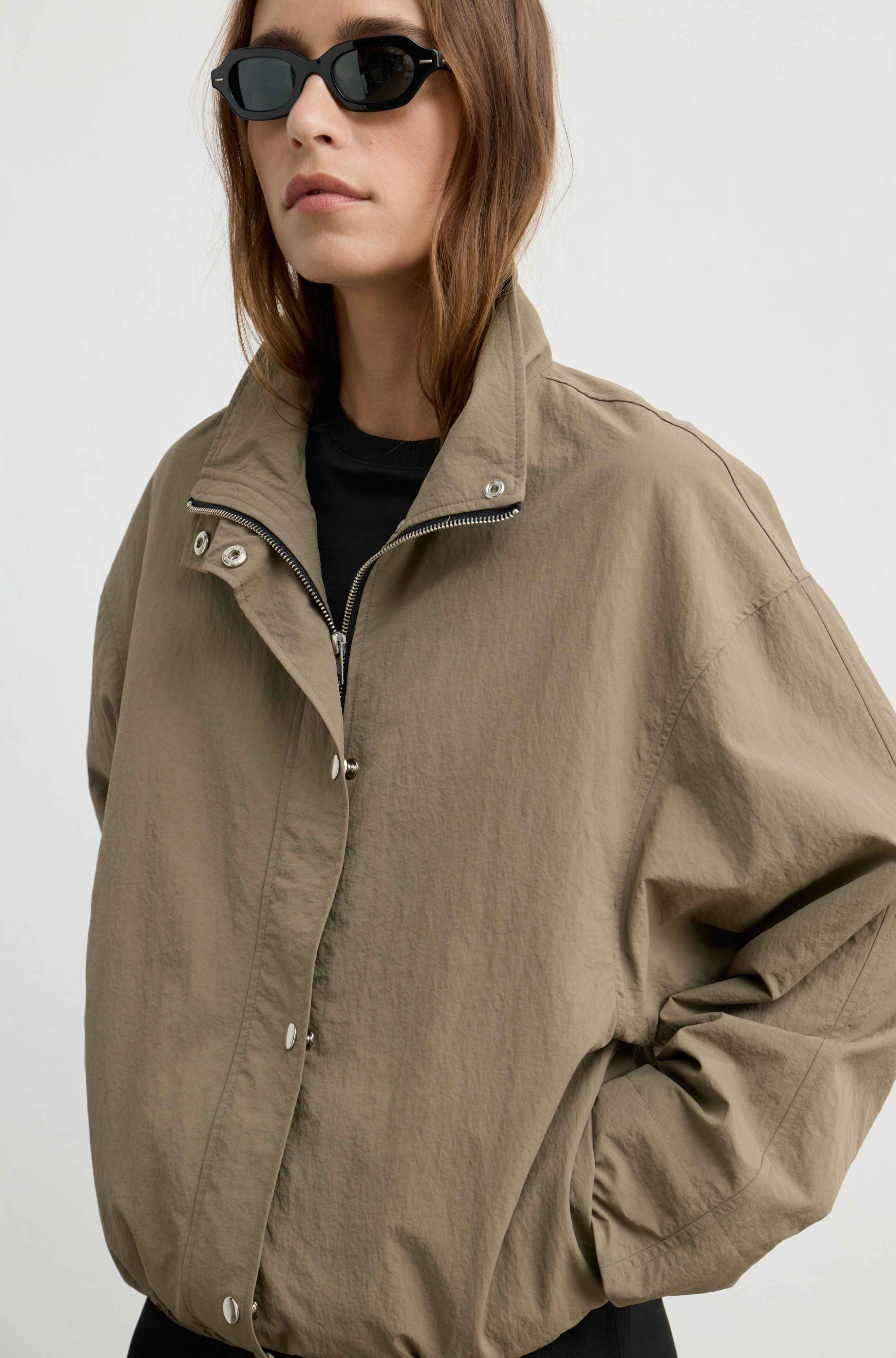 A woman wearing a khaki utility jacket with a high collar and silver snap buttons over a black top, styled with black sunglasses, against a plain light gray background.