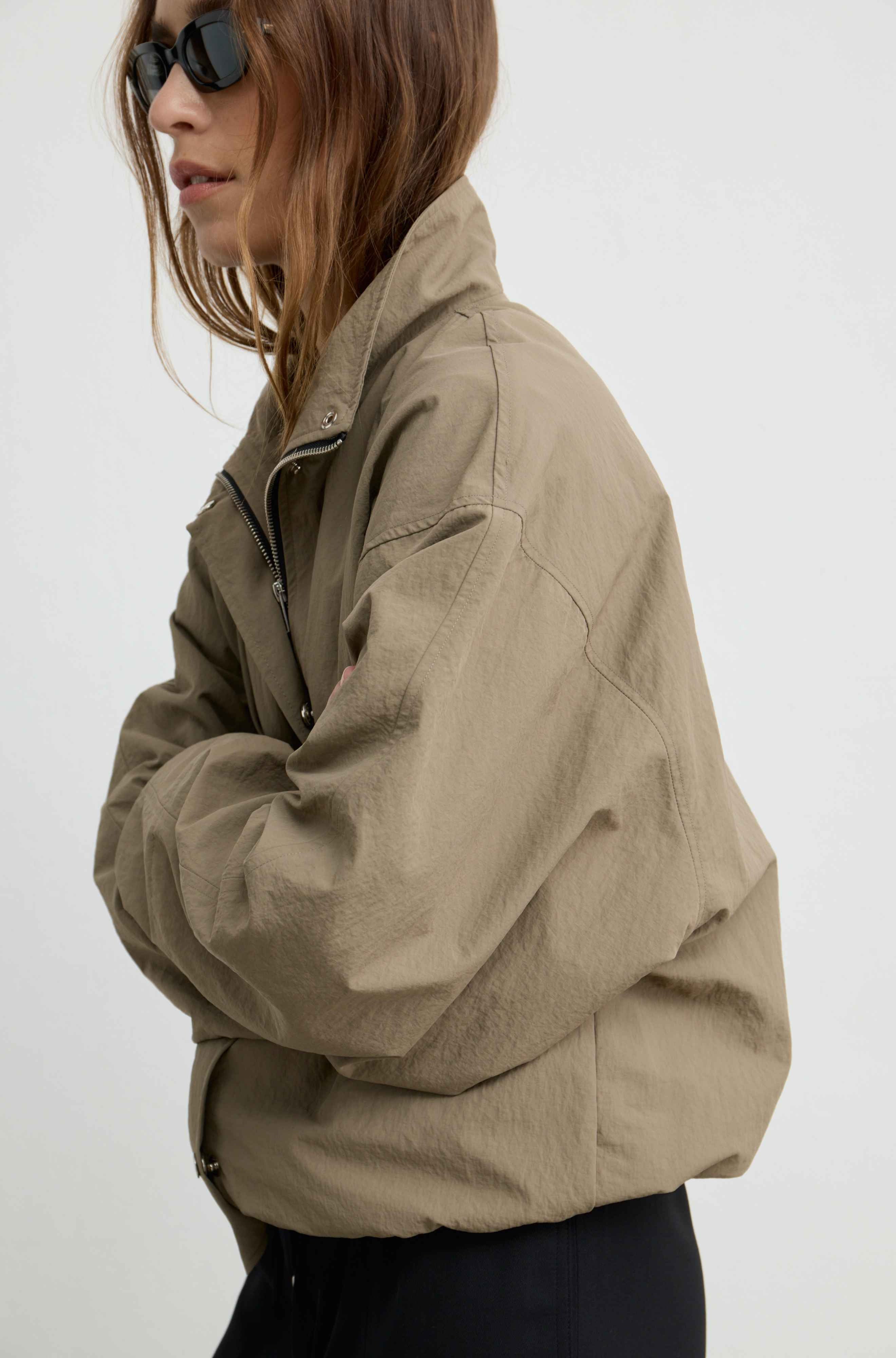 A woman wearing a cropped, oversized utility jacket in a warm khaki color, styled with black pants and dark sunglasses against a plain white background.