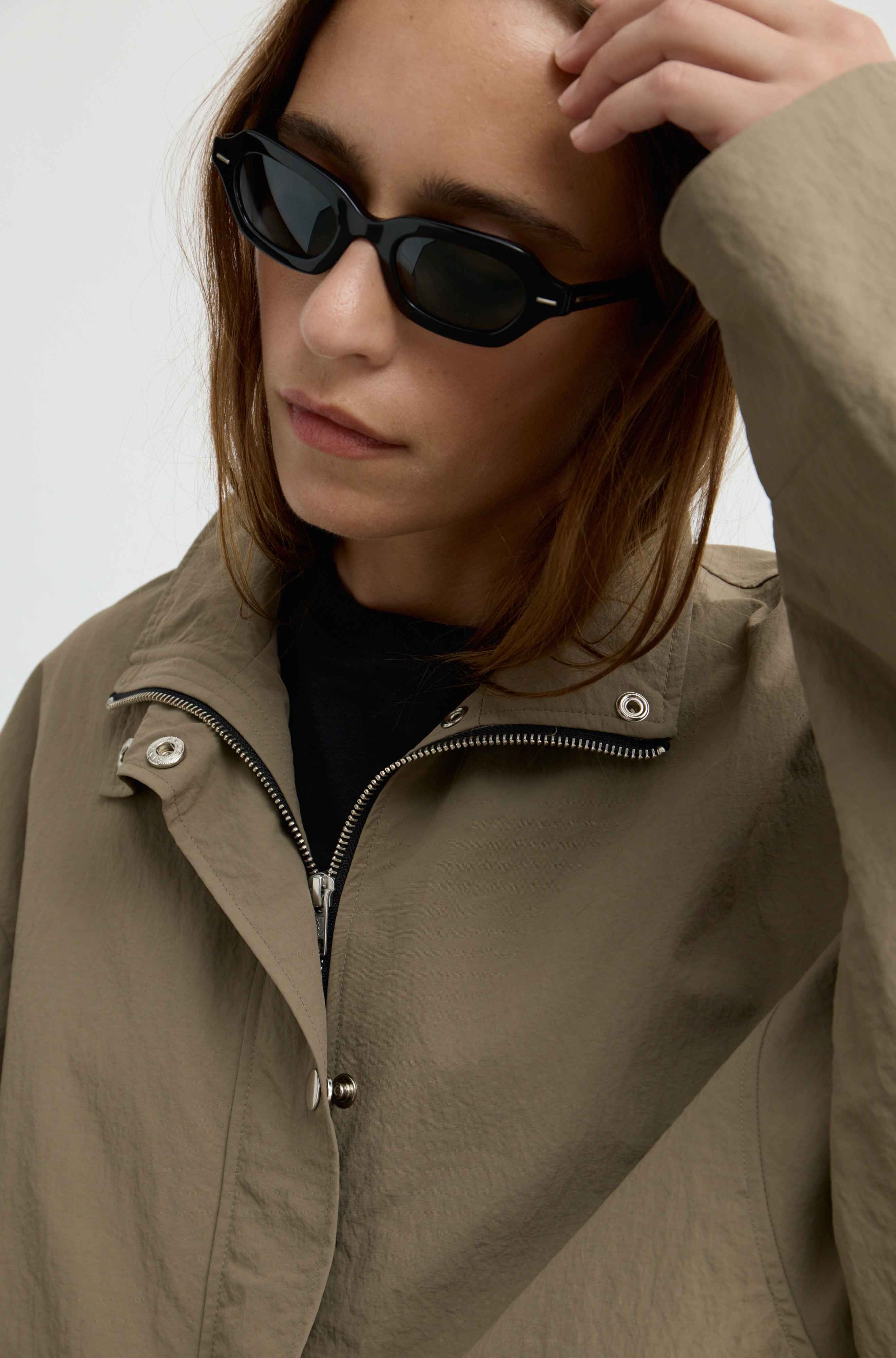 A close-up of a model wearing black cat-eye sunglasses and a khaki utility jacket over a black top, against a plain light background.