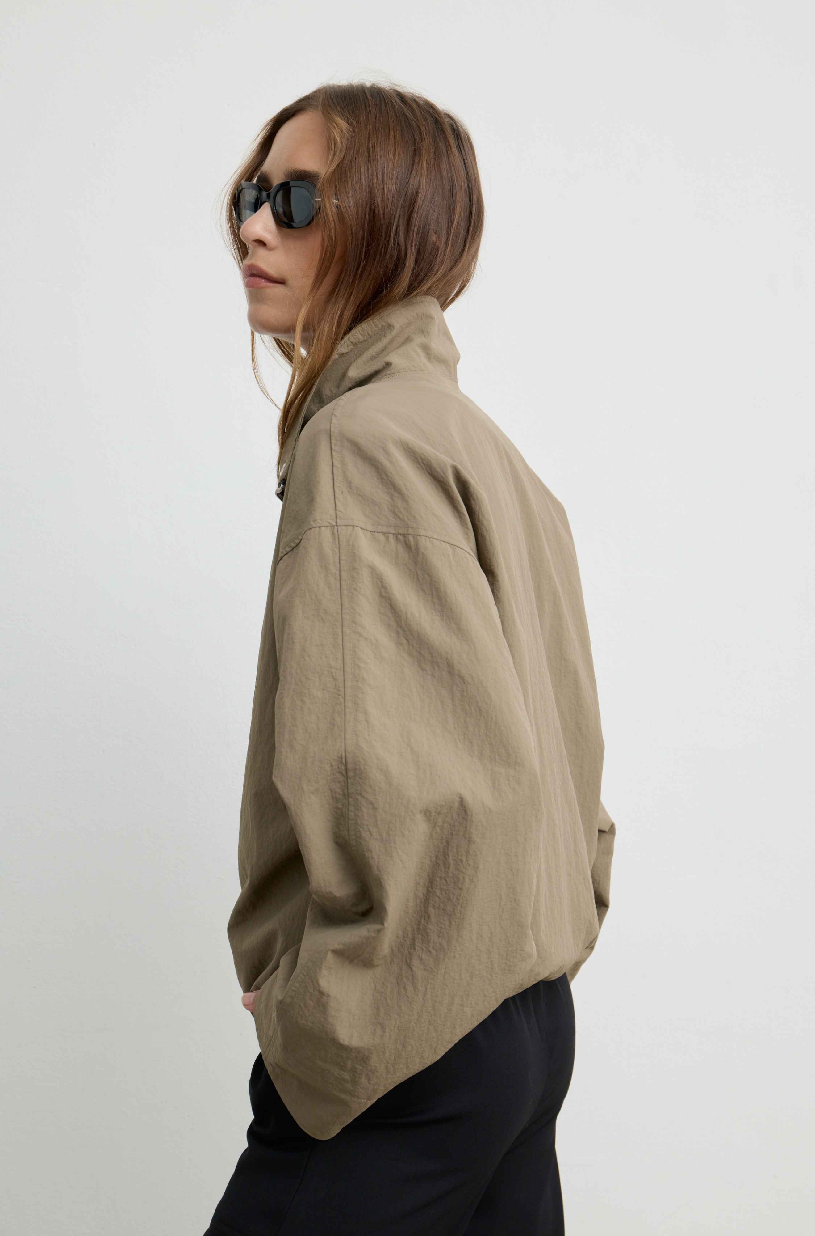 A woman wearing a light brown high-collar jacket and dark pants, shown from a side profile against a plain light gray background.