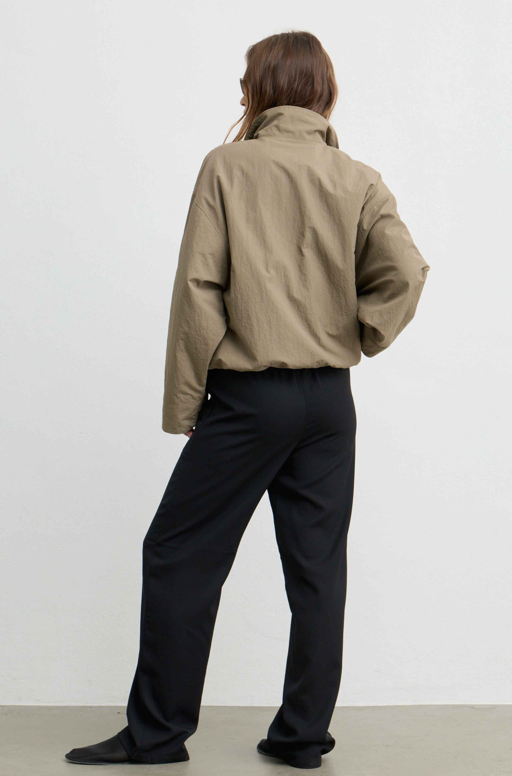 A model shown from the back wearing a cropped, khaki utility jacket and black trousers, standing against a plain white studio wall.