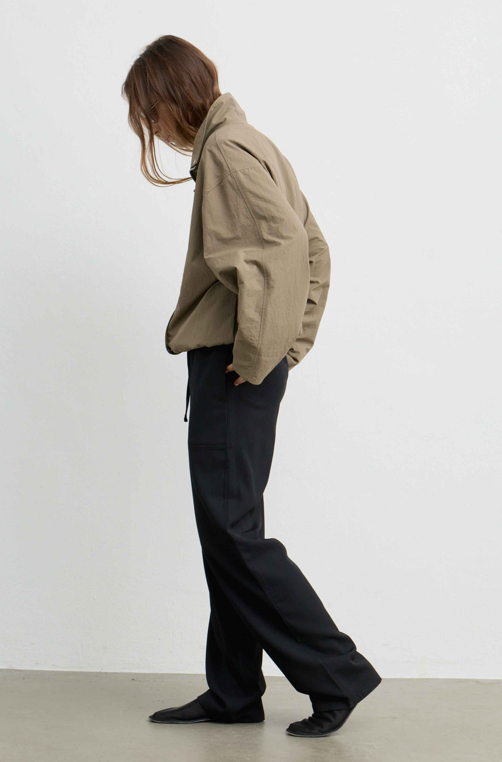 A model wearing a khaki cropped utility jacket and black wide-leg trousers, shown from a side profile against a plain white studio wall.