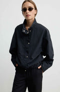 A model wearing a black lightweight jacket with a high collar and snap details, paired with black trousers and dark sunglasses, against a plain light gray background.