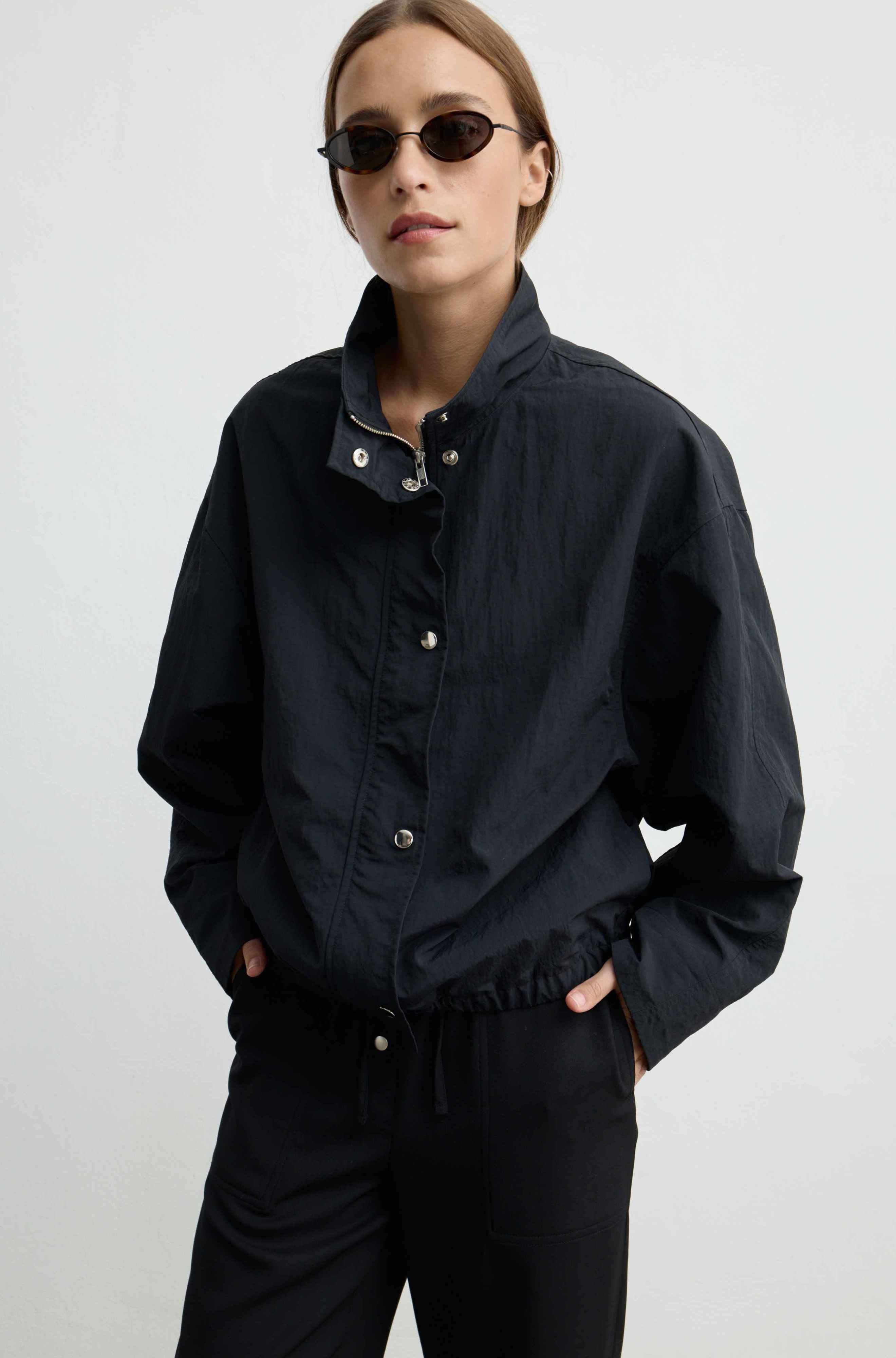 A model wearing a black lightweight jacket with a high collar and snap details, paired with black trousers and dark sunglasses, against a plain light gray background.