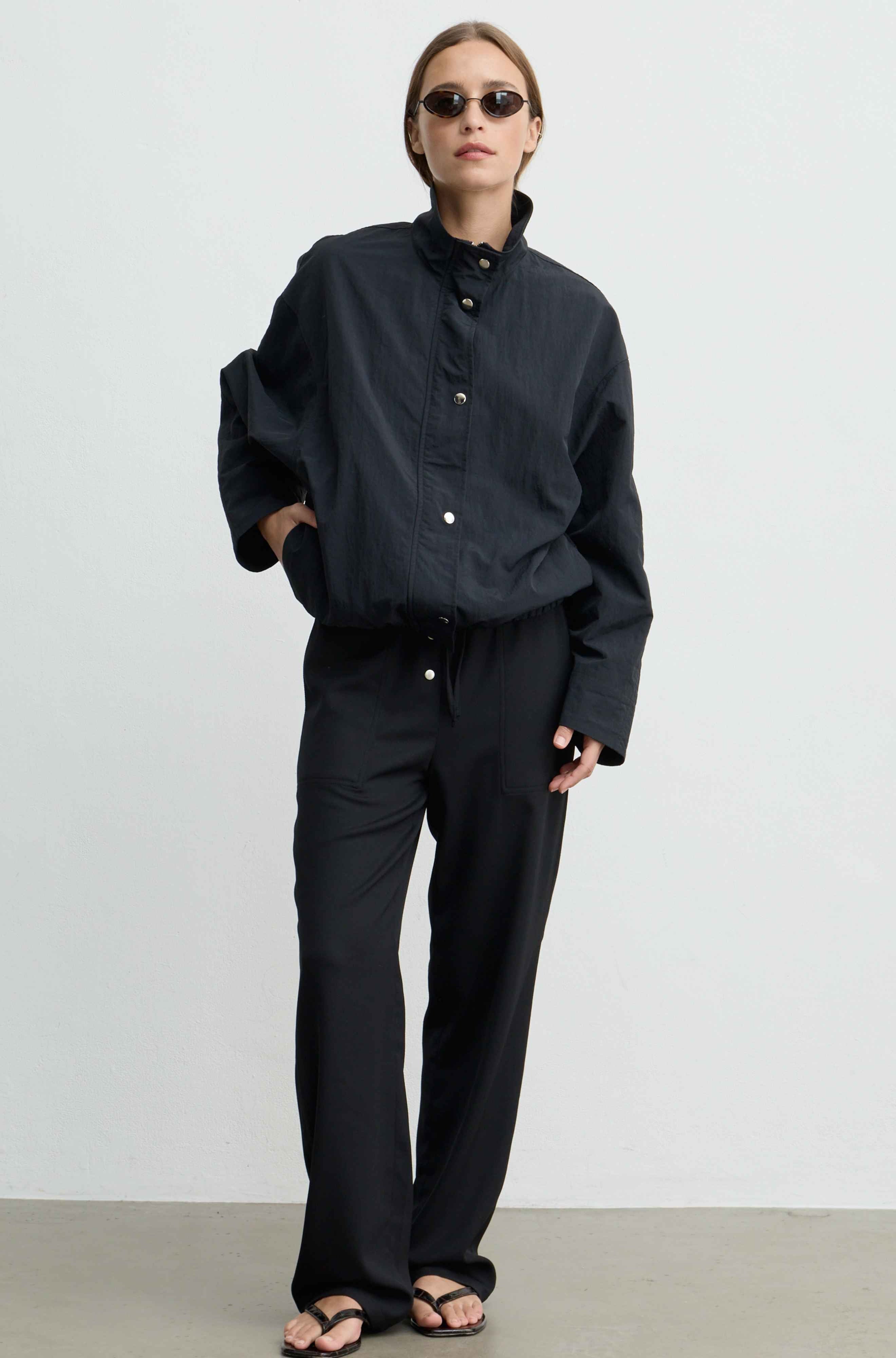A model wearing a matching black jacket and black button up pants, styled with black sunglasses and black sandals against a plain light gray studio background.