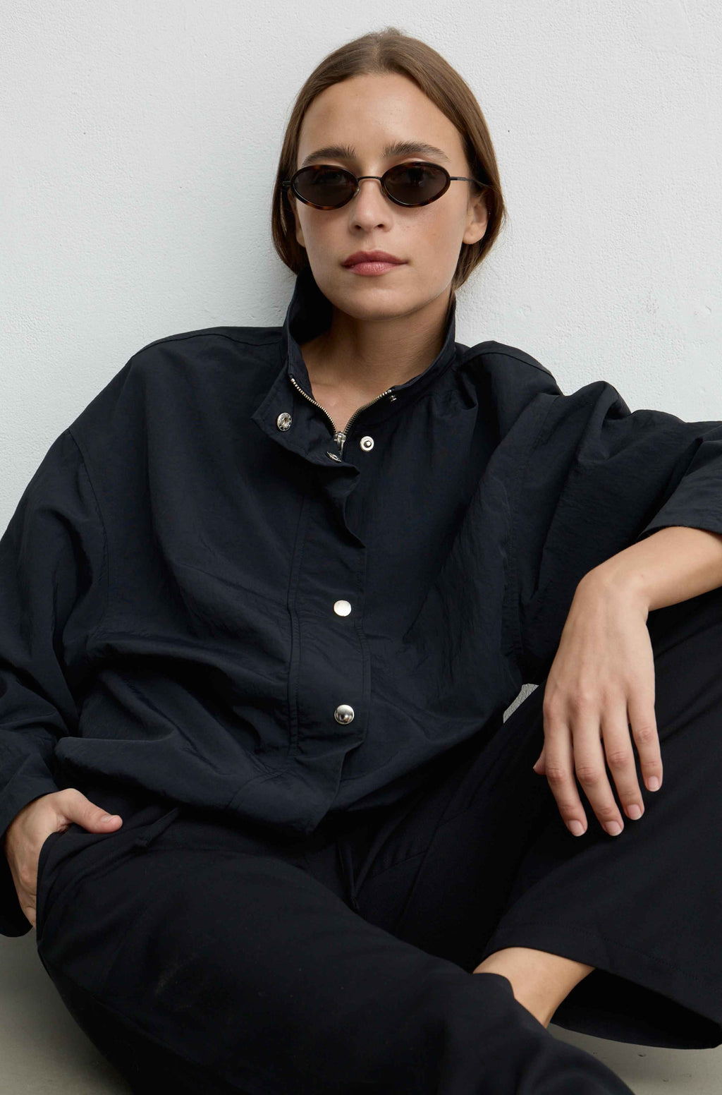 A woman wearing a loose black button-up jacket and black pants, seated against a light gray wall. She is wearing small dark sunglasses, and the photo is styled in a clean, minimalist setting.