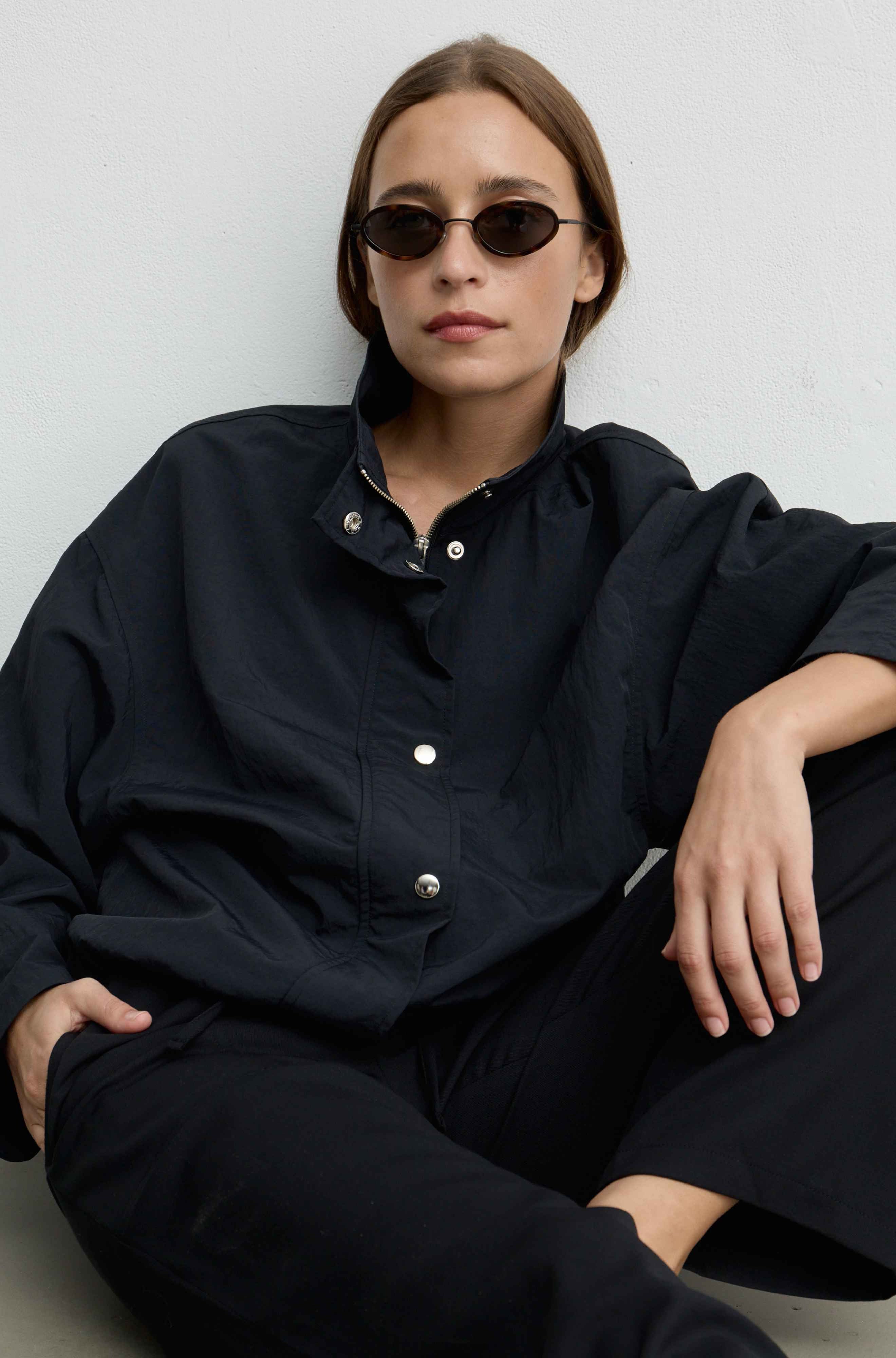 A woman wearing a loose black button-up jacket and black pants, seated against a light gray wall. She is wearing small dark sunglasses, and the photo is styled in a clean, minimalist setting.