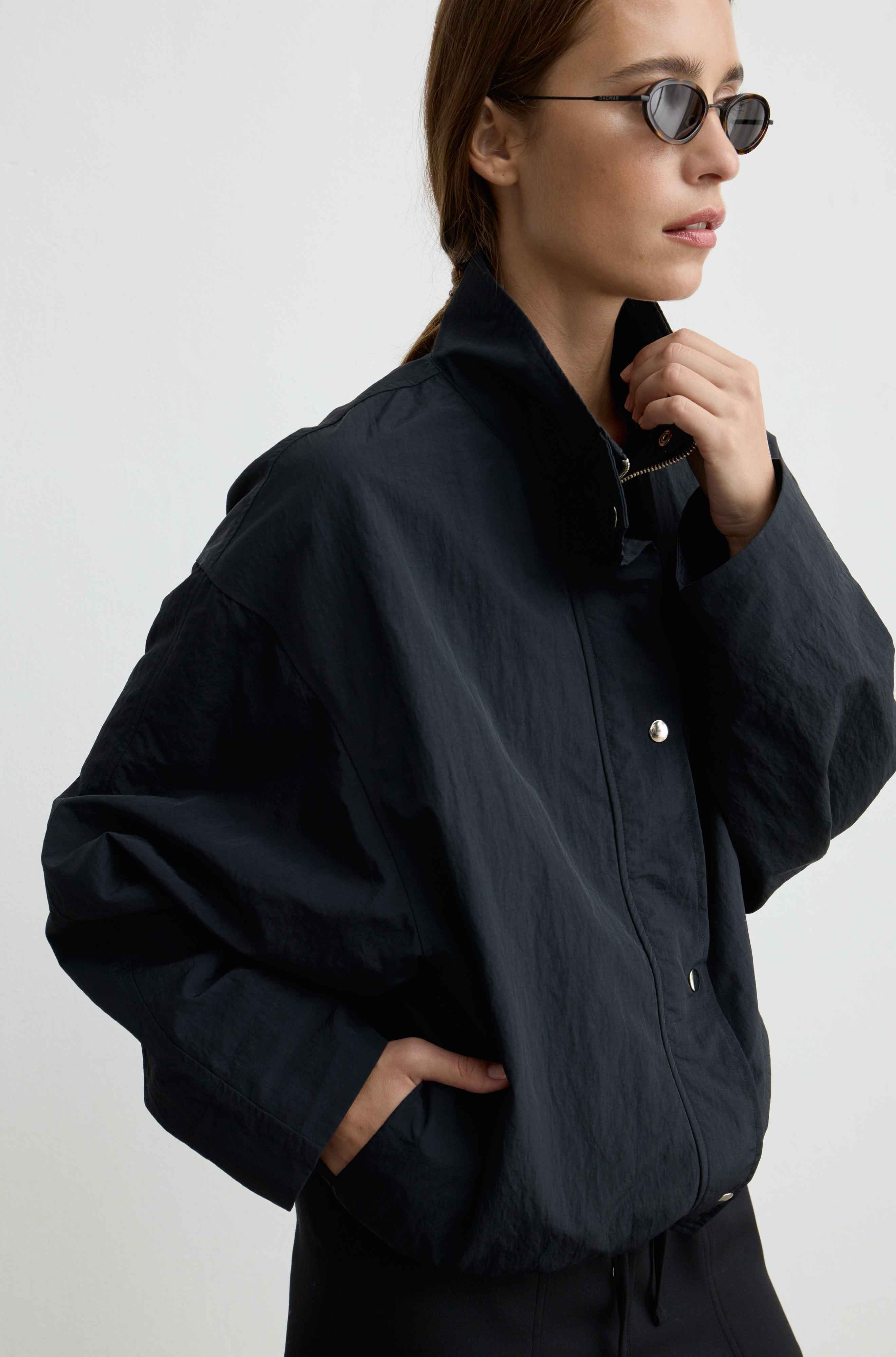 A woman wearing a black lightweight jacket with a high collar and silver snap buttons, styled with black pants and sunglasses against a light gray background.