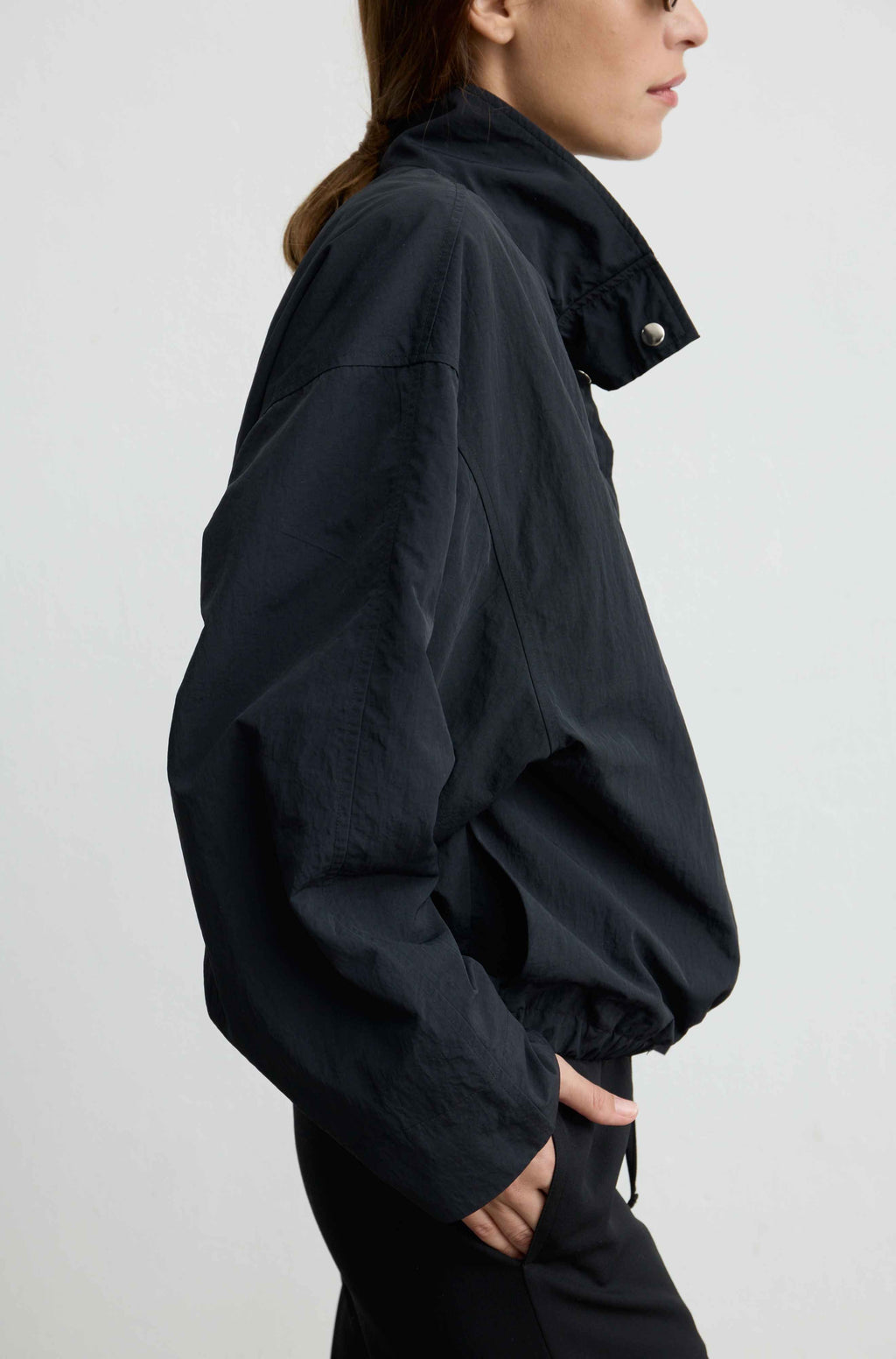 A side-profile view of a model wearing a black lightweight jacket with a high collar and silver snap button, paired with black pants, against a plain light gray background.