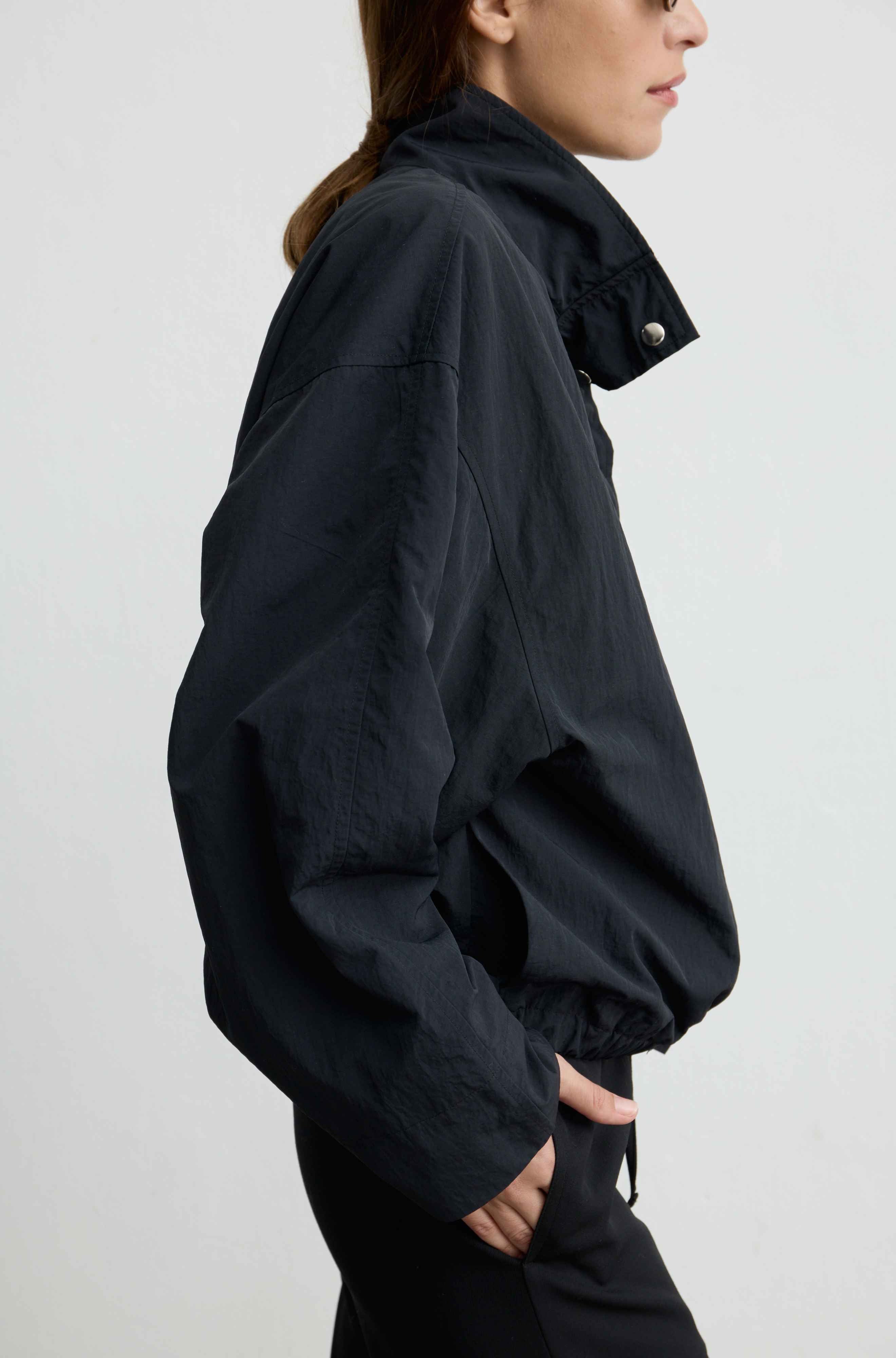 A side-profile view of a model wearing a black lightweight jacket with a high collar and silver snap button, paired with black pants, against a plain light gray background.