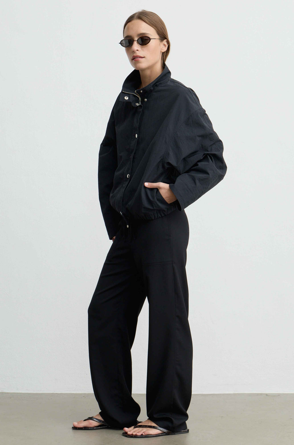 A model wearing a black lightweight jacket and matching wide-leg trousers, styled with black sandals and sunglasses, standing against a plain light gray studio background.