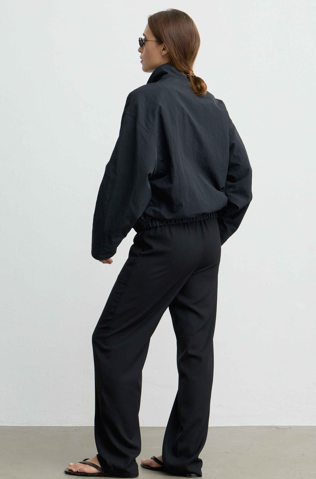 A woman wearing a black lightweight jacket and matching black pants, photographed from the back in a minimalist studio setting with a white wall and light gray floor.